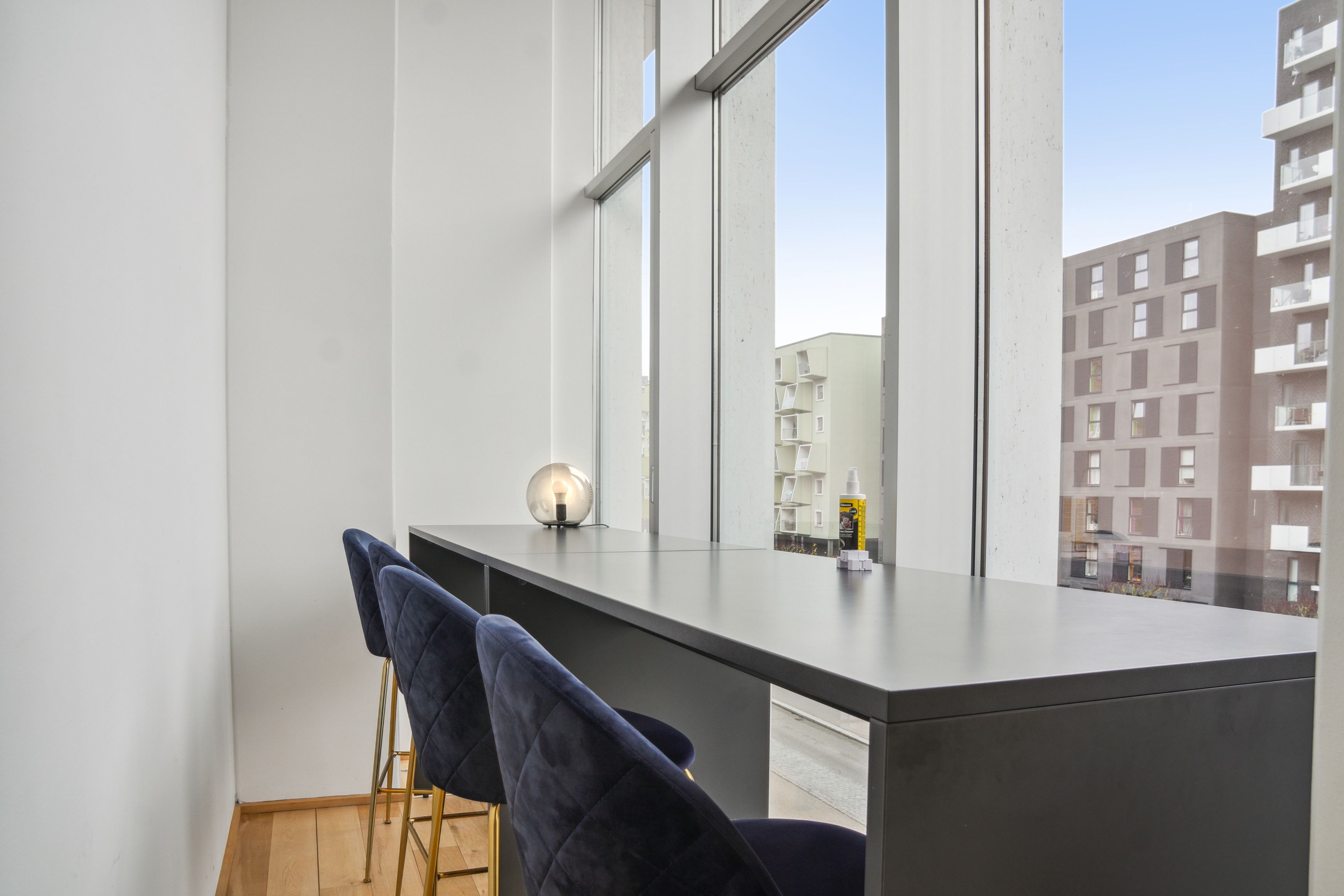 Moderne kontor med lyse, store vinduer og smukke trægulve. Et langt bord med fire blå stole og en lampe. Bygninger i baggrunden og en dejlig udsigt til haven.