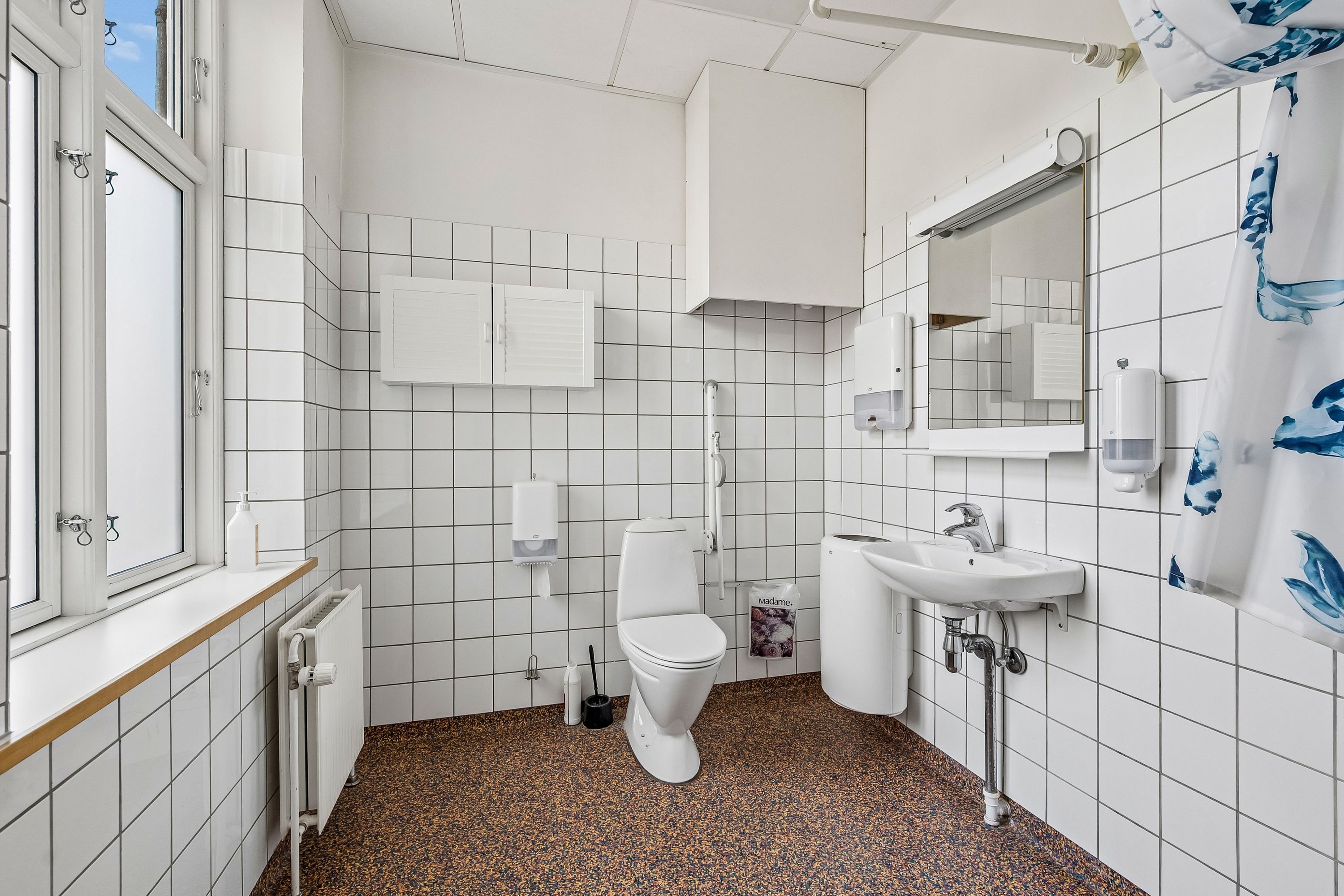 Kontor med hvidt flisebelægning, toilet og håndvask. På væggen er der flere håndsæber og en håndklædeholder. På gulvet er der brungul gulvbelægning. Vinduet med rammer er åbent og ud til gaden