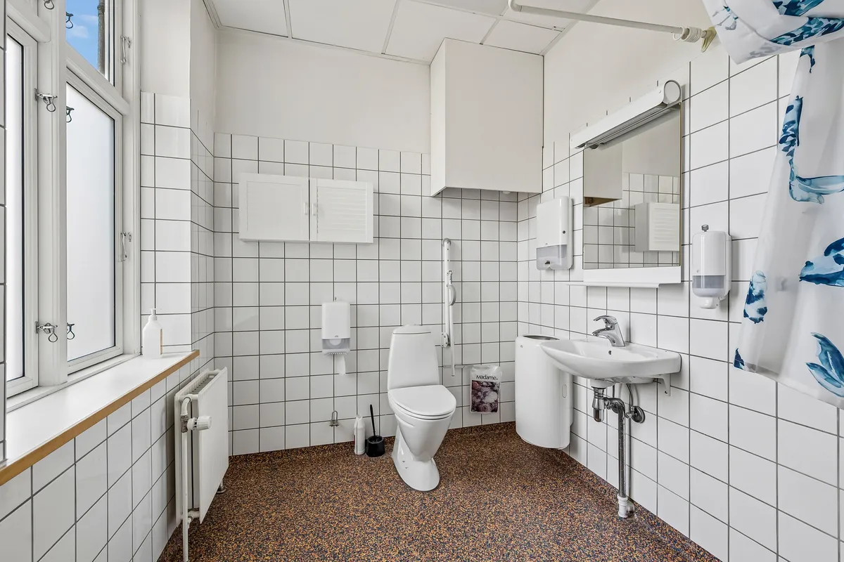 Kontor med hvidt flisebelægning, toilet og håndvask. På væggen er der flere håndsæber og en håndklædeholder. På gulvet er der brungul gulvbelægning. Vinduet med rammer er åbent og ud til gaden