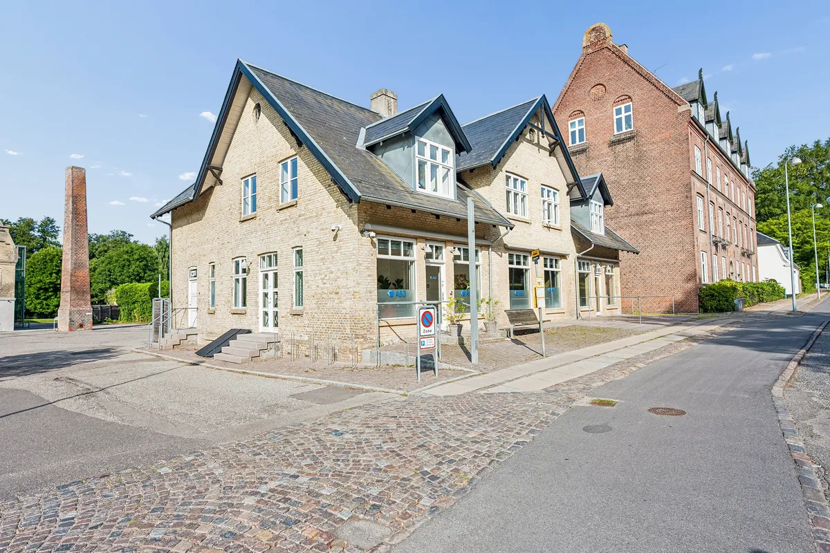 Kontorby i Lyngby med røde mursten og stenbro foran hoveddøren. En høj skorsten og et rødt tegltag ses i baggrunden. En søjle med skilt og grønne planter.