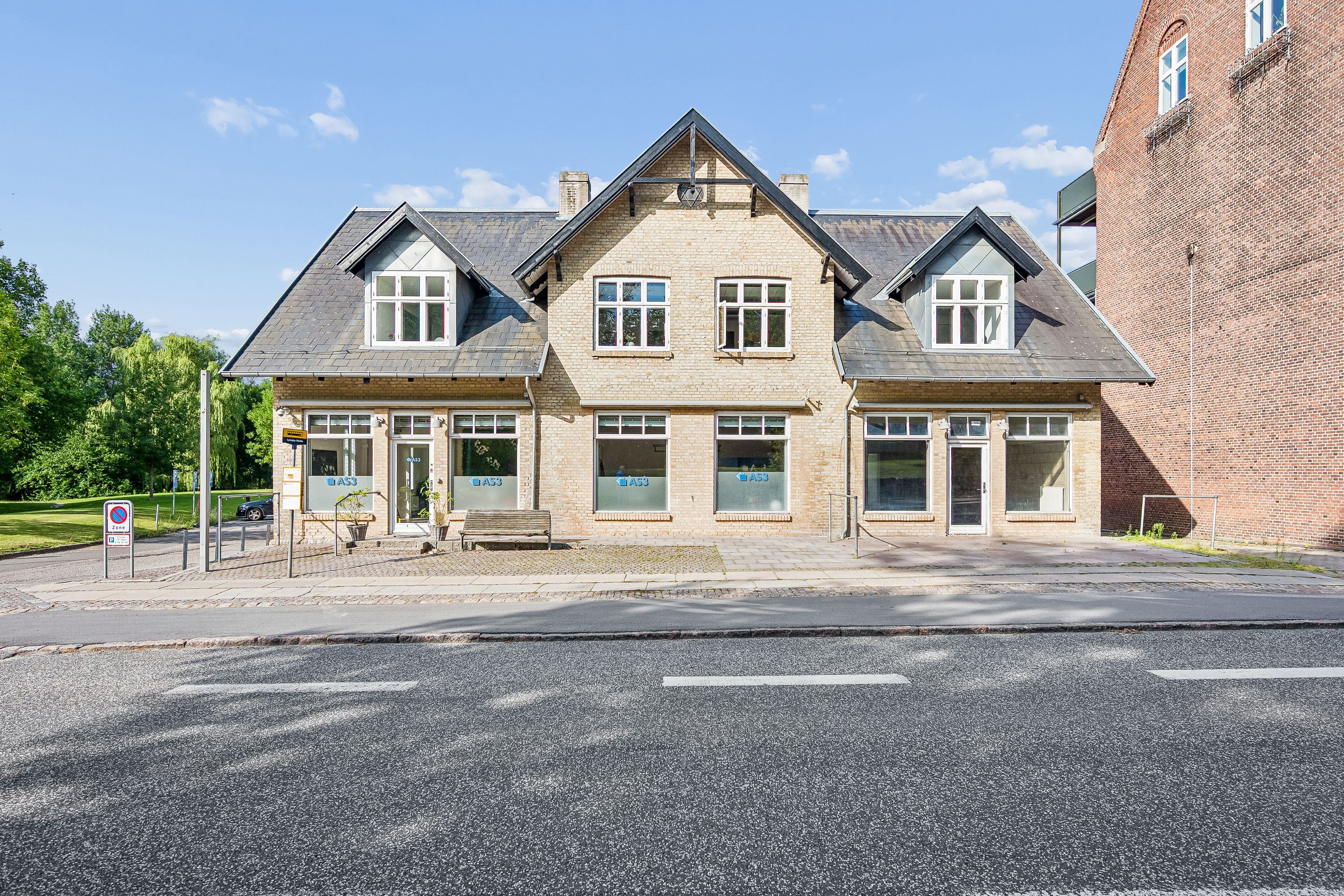 Butikslokaler med store vinduer og slanke facader i Kongens Lyngby. En parkeringsplads med parkeringsforbudsskilt og grønne områder ligger foran bygningen.