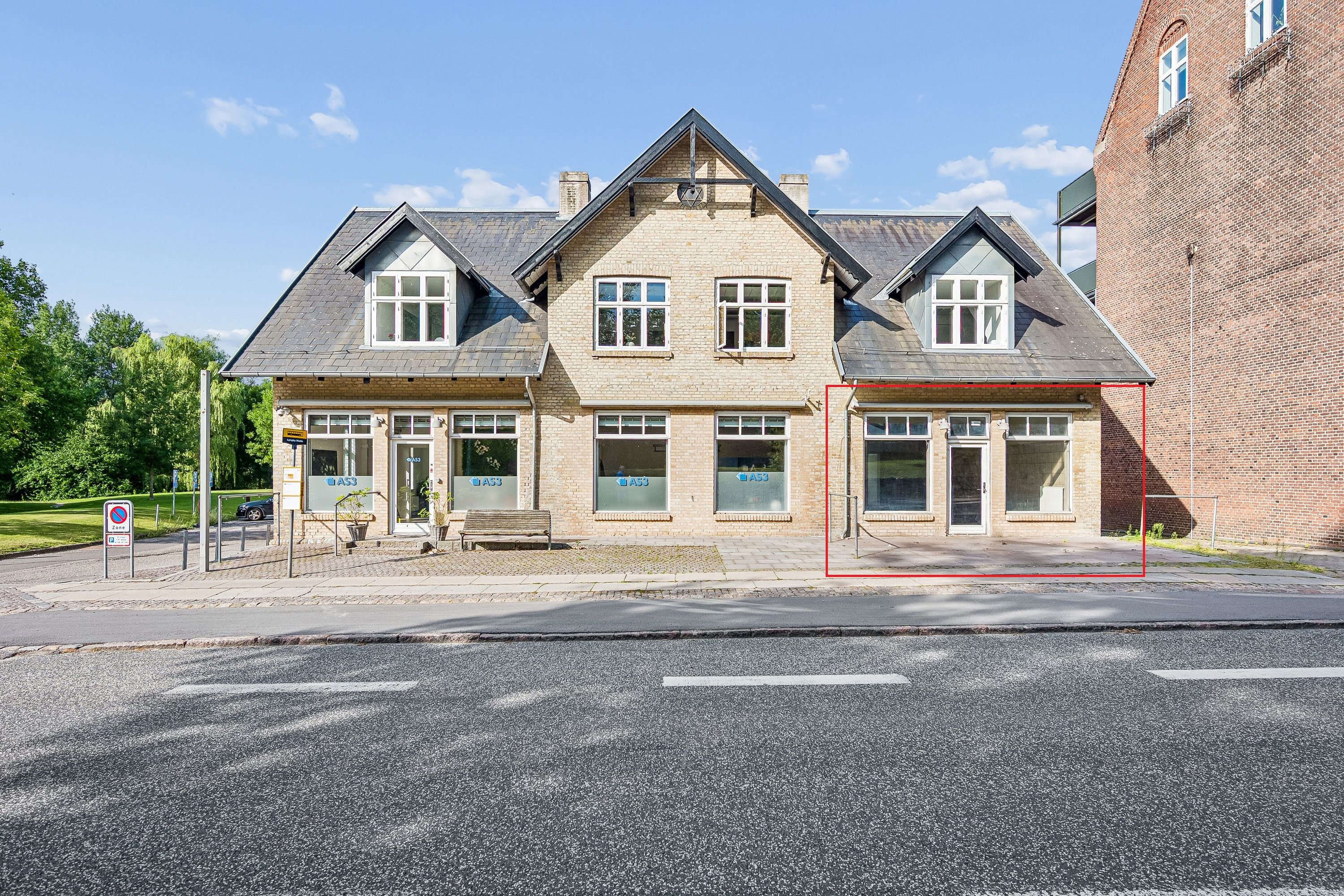 En stor butik med mærke AS3 i Lyngby Hovedgade 10B, 2800 Kongens Lyngby. Butikken med røde kantlinjer har to vinduer og en indgang. Parkeringsforbudsskilt og gul vejvisningsskilt står på fortovet