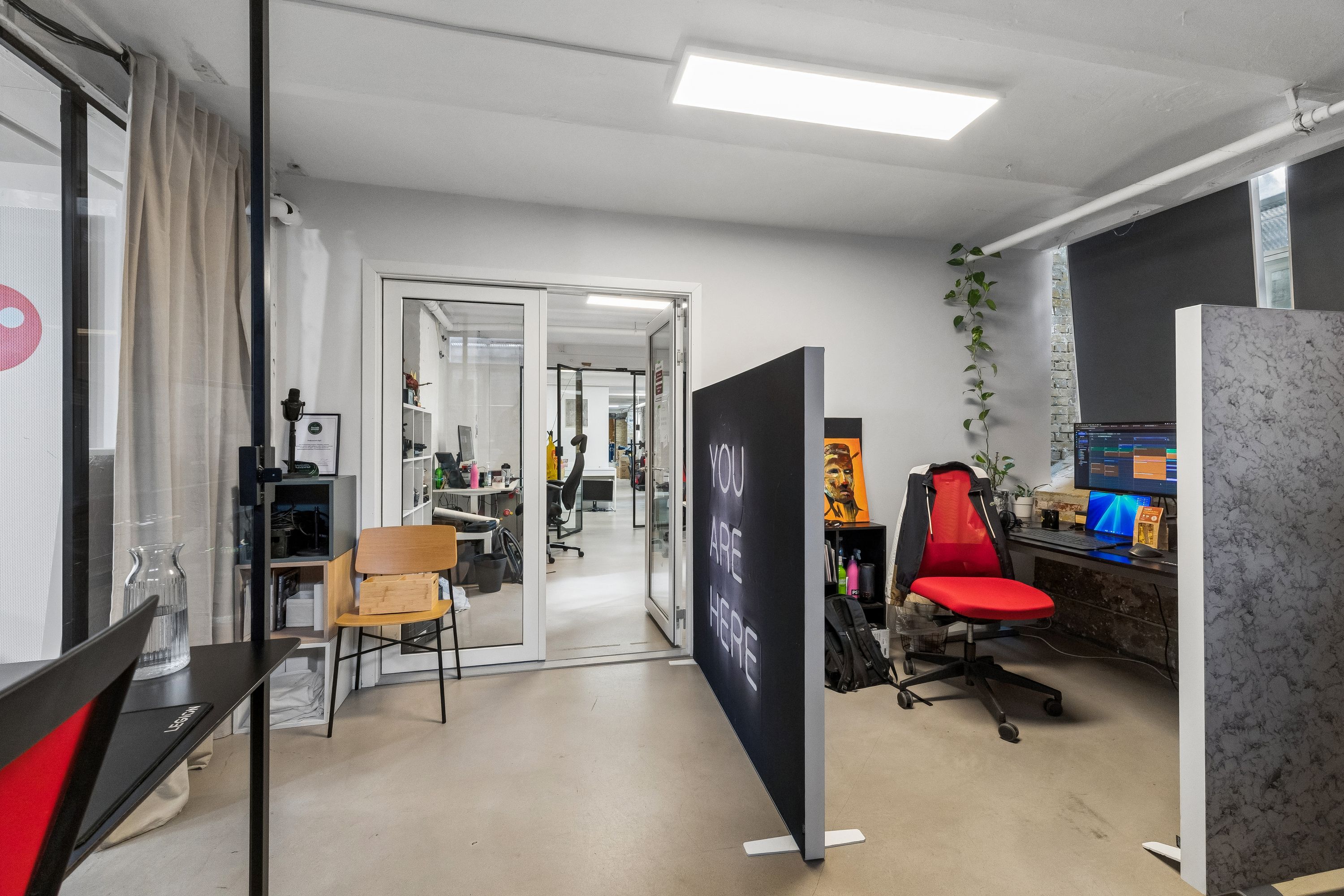 Et moderne kontor med glasdøre, højt gulv og en stor skilt med teksten You are here. I rummet er der en skrivebord med computer og to stole samt et plante og lysekroner