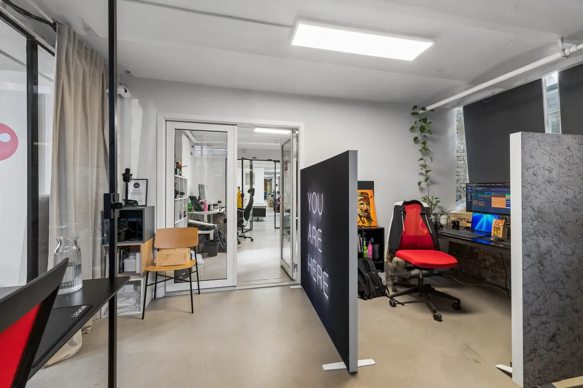 Et moderne kontor med glasdøre, højt gulv og en stor skilt med teksten You are here. I rummet er der en skrivebord med computer og to stole samt et plante og lysekroner