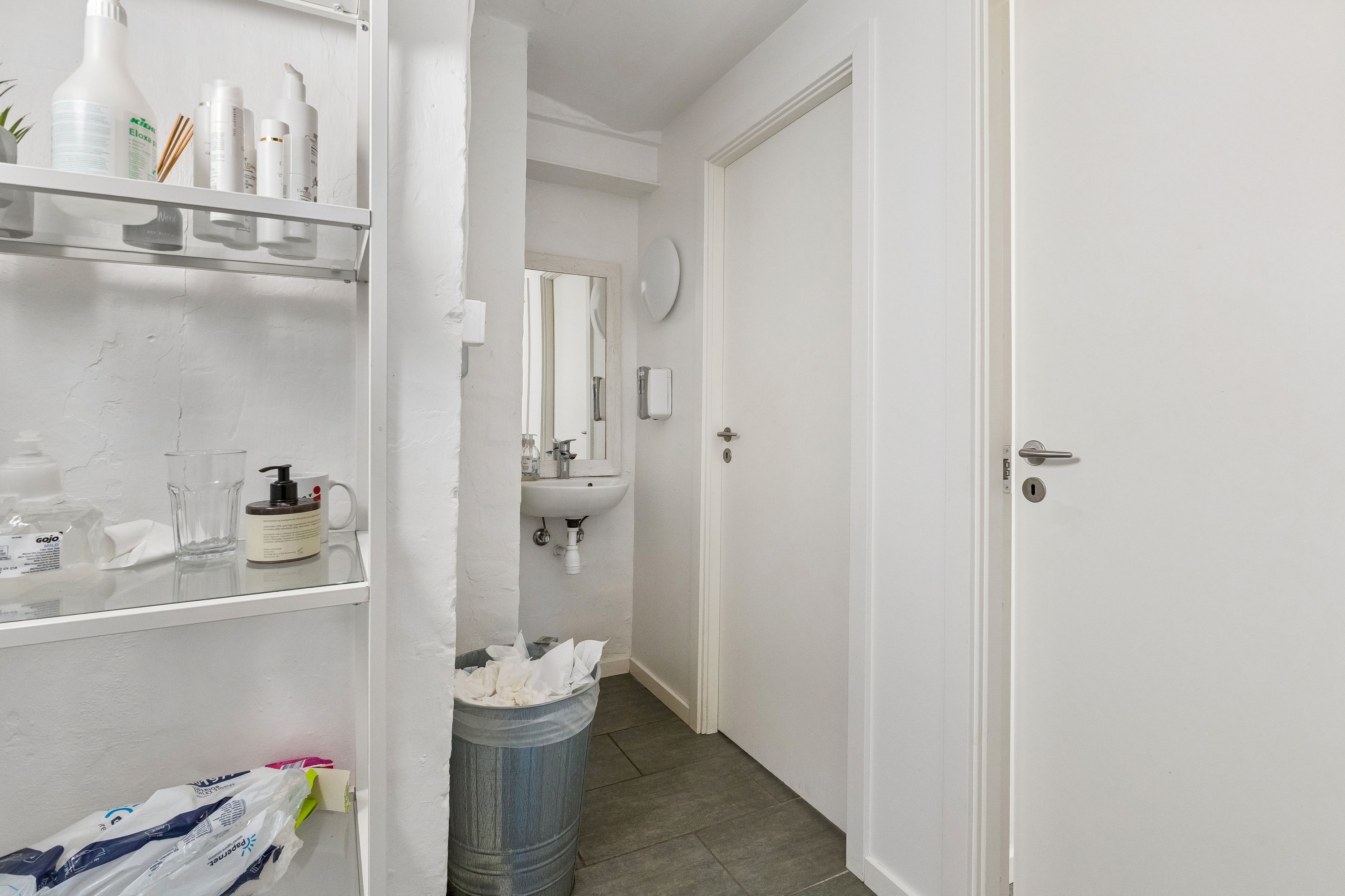 Rent, rent toilet med håndvask og spejl, opbevaringsskabe med kosmetik og toiletartikler samt papposer og skraldespand i gulvet