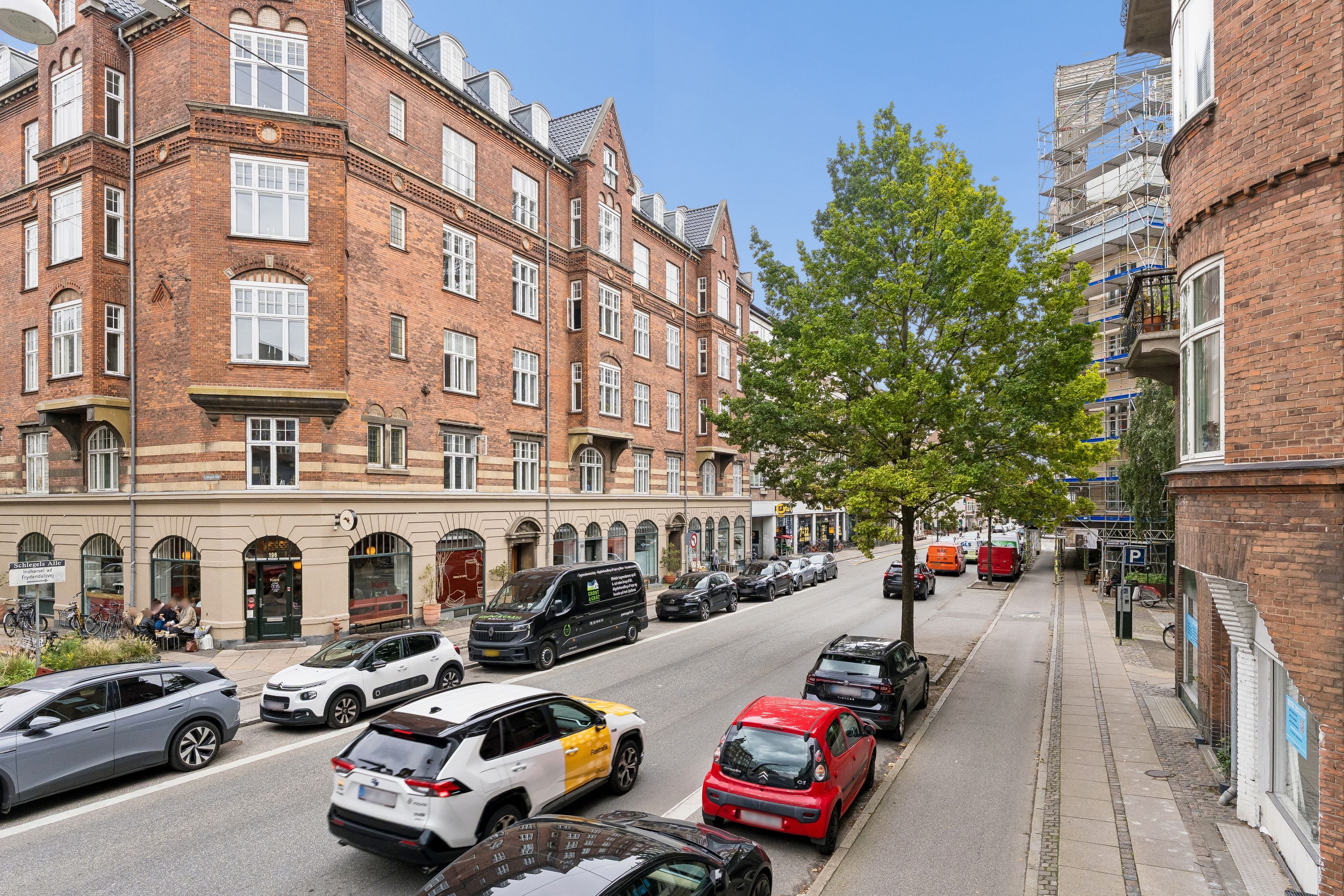 Parkerede biler og bygninger langs Vesterbrogade, hvor det er muligt at leje kontorer og butikker. Høj synlighed på gaden med facadevinduer i de røde og gule bygninger.