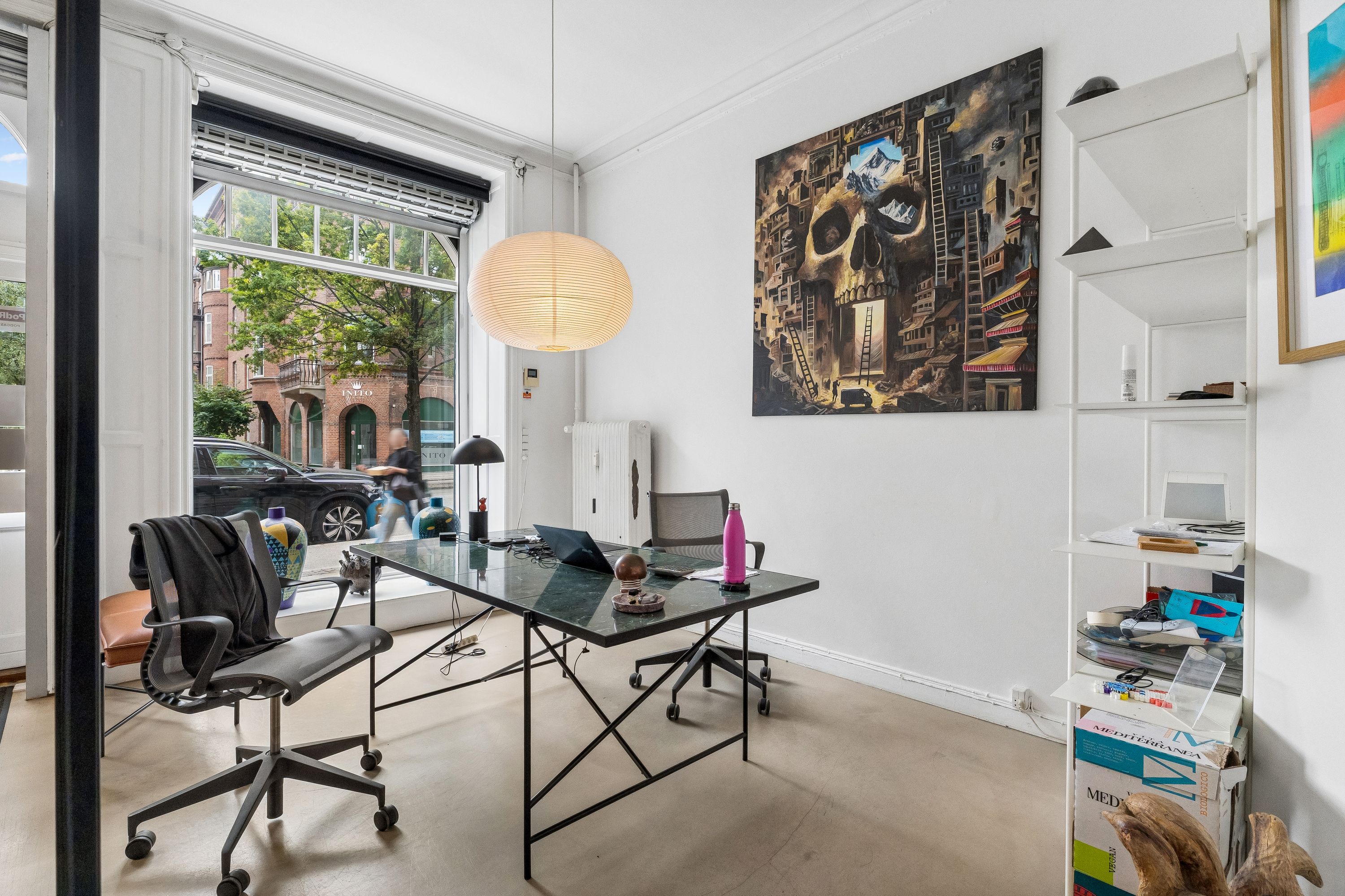Kontor med moderne møbler, skrivebord med laptop, køkkenbord, stol, lamper og store ruder med udsigt til gaden.