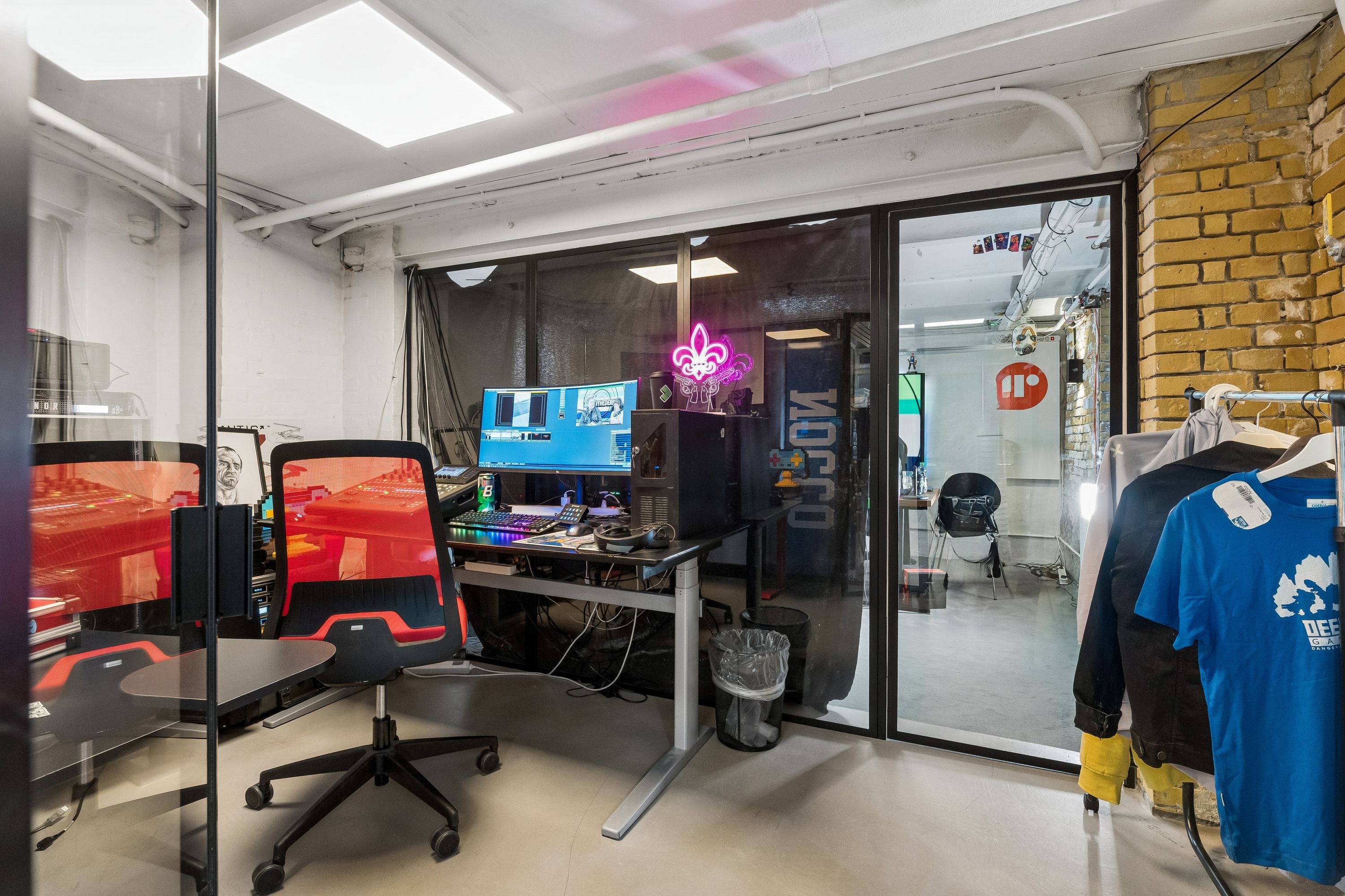 Kontor med flere skriveborde og stole samt tøj på tøjstativ og neonskilt med teksten "MOCO"