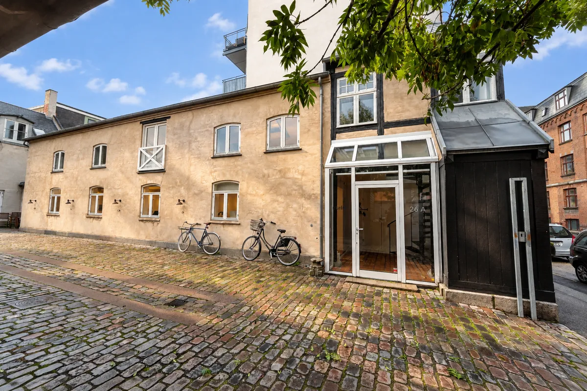 To cykler står parkeret foran et bindingsværkshus. Bygningen er gulmalet med vinduer og en stor glasdør. Stiger og facader er pænt vedligeholdt