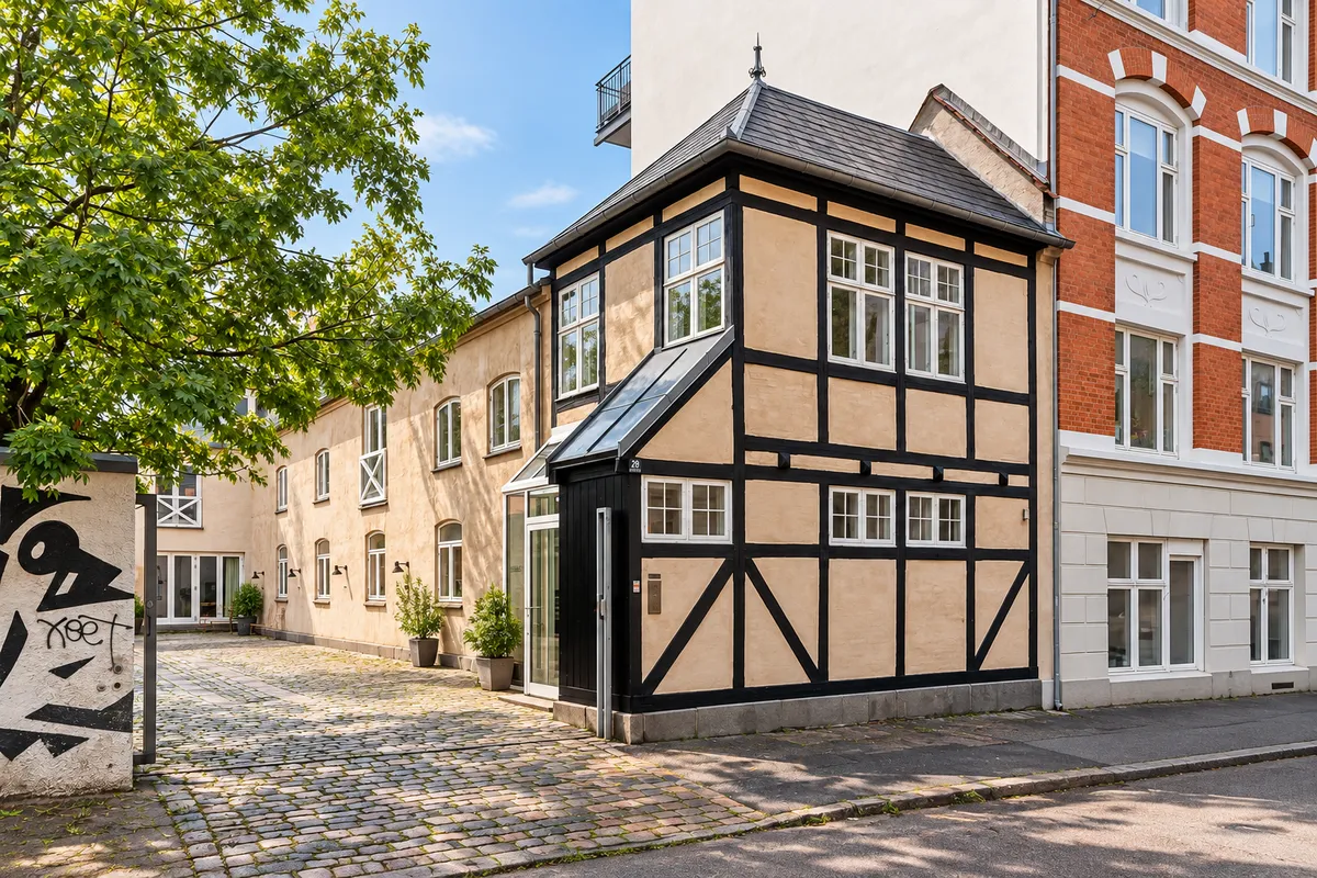 Lyst kontormiljø med træ og tøj på Svanevej 26A. Bindingsværkshus med lyse, pæne kontorer. Planter og træer foran huset. Cobblestone vej med grafitti på siden