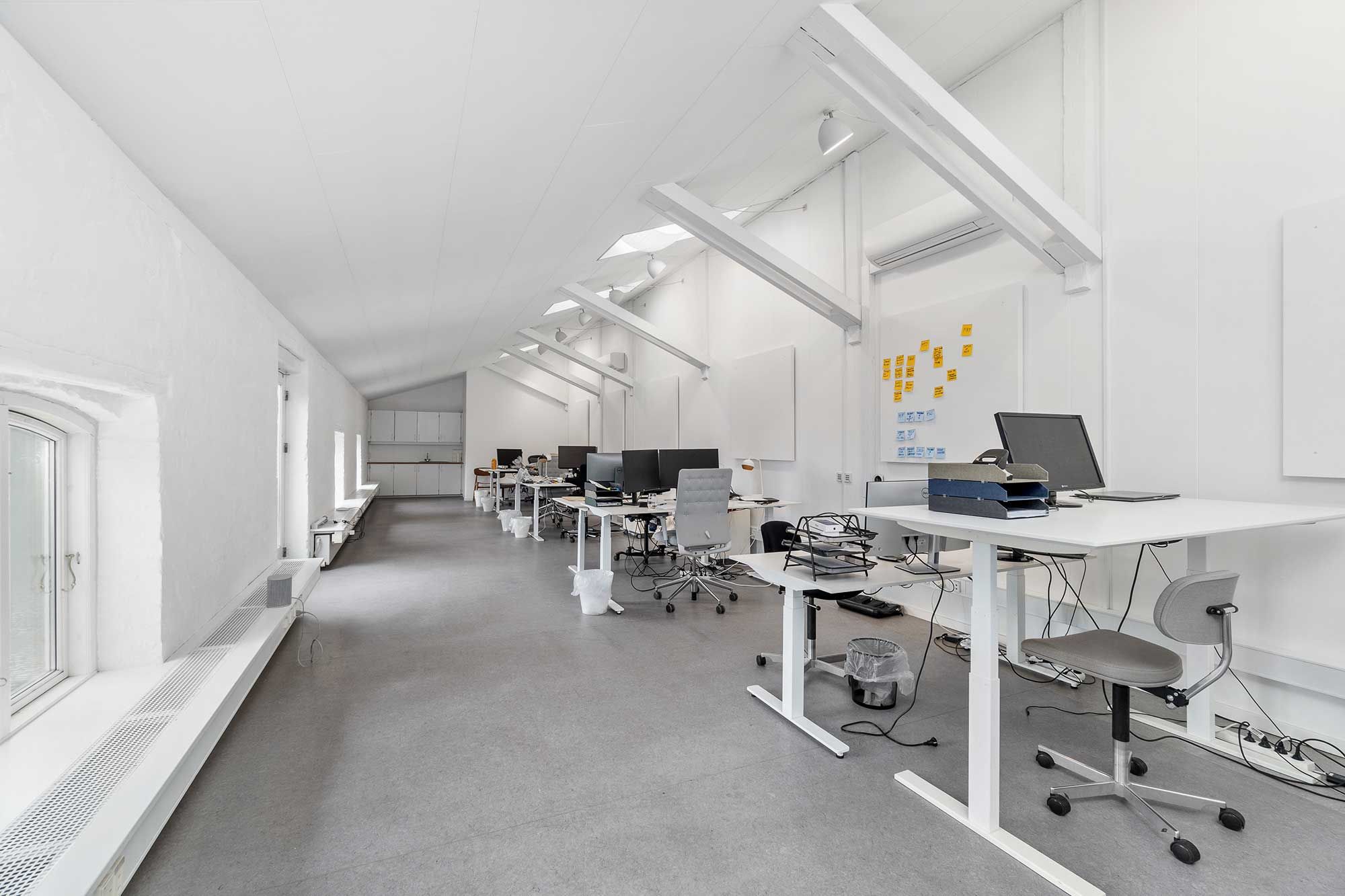 Kontor med højt til loftet, hvidt loft, gulve, skriveborde, computere, skrivebordslys, stolpe og luftkanaler. Mange skriveborde er placeret i en række.