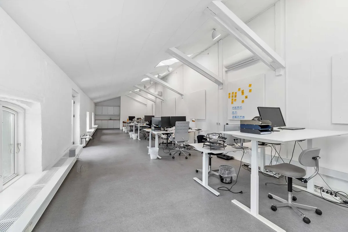 Kontor med højt til loftet, hvidt loft, gulve, skriveborde, computere, skrivebordslys, stolpe og luftkanaler. Mange skriveborde er placeret i en række.