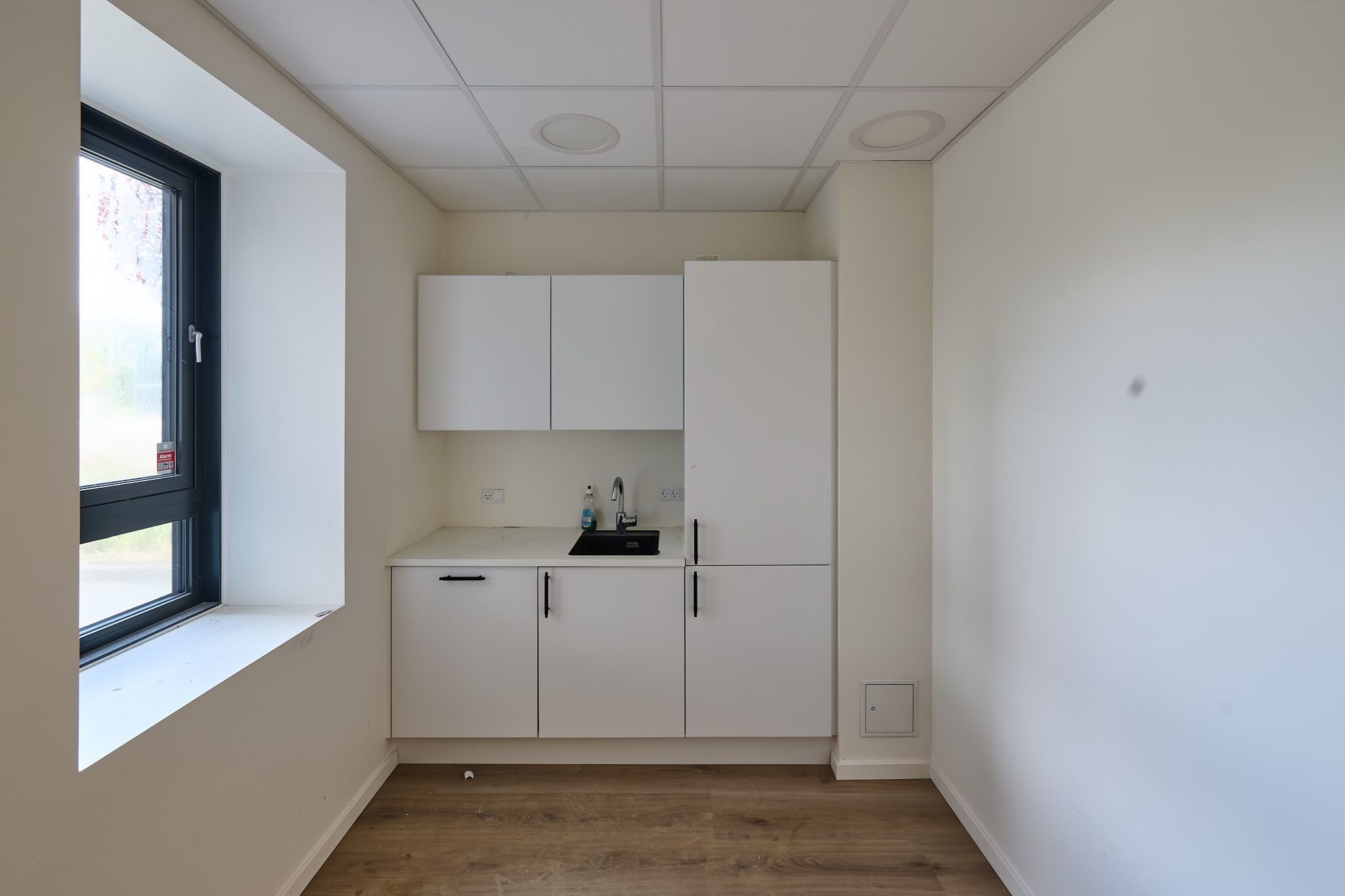 Et lille køkken med hvidt loft og hvide vægge, hvor der er en hvid skab samt en høj kabinet. Et vindueskarmen med et glasvindue og udsigt til grønne træer.