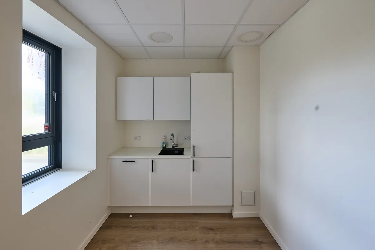 Et lille køkken med hvidt loft og hvide vægge, hvor der er en hvid skab samt en høj kabinet. Et vindueskarmen med et glasvindue og udsigt til grønne træer.