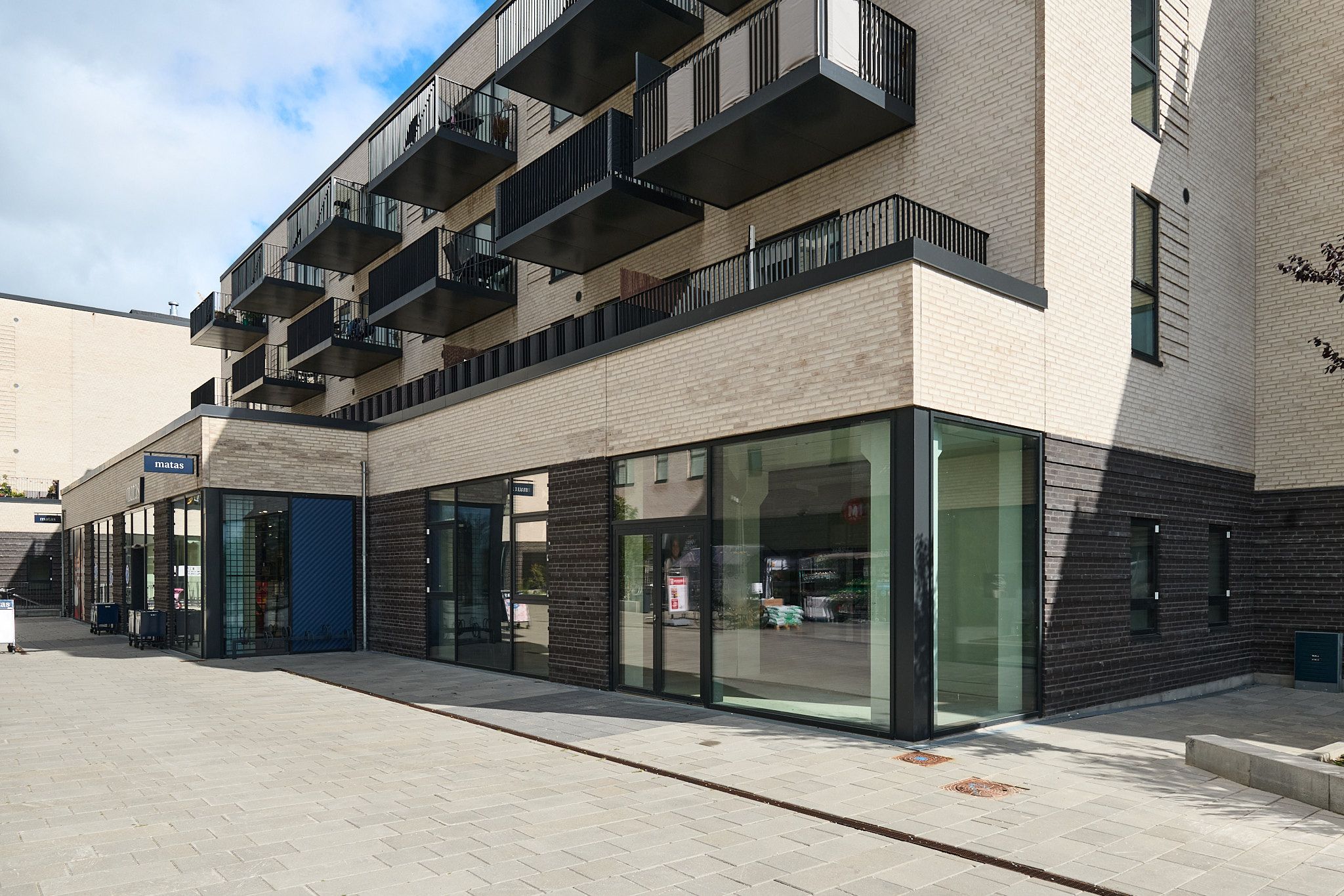 Et moderne og velplejet lejemål med glasvinduer og terrasser på 2. og 3. etage. Balkonner og terrasser er udstyret med mørke stålrækværk. På jorden er der en gulvbelægning af lysegrå og grønne tegl.