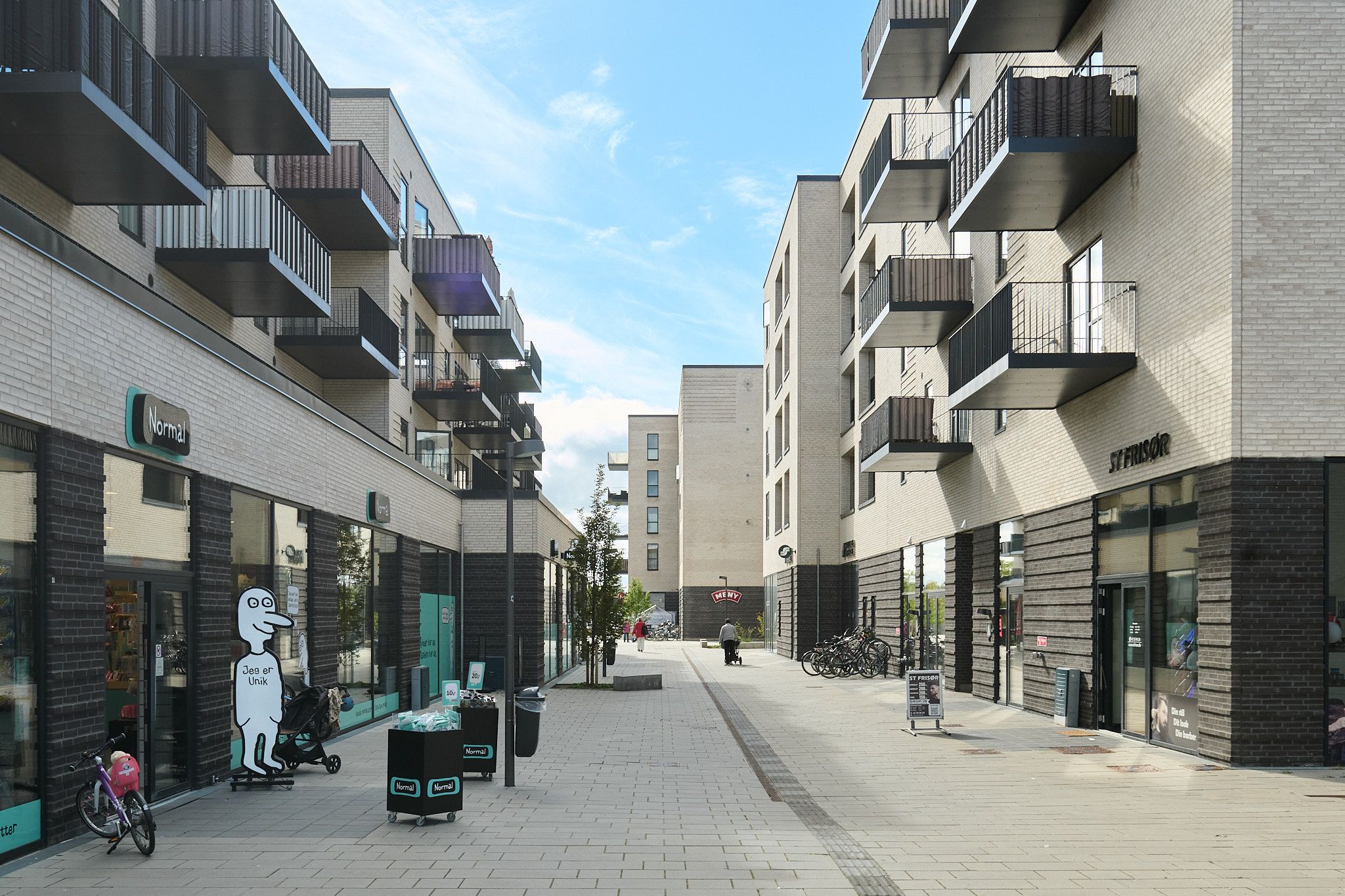 Lejlighedsbygninger med balkoner og solpaneler og torve med butikker og stående cykler på Torvestrædet i Skovlunde. En person går med en babyvogn.