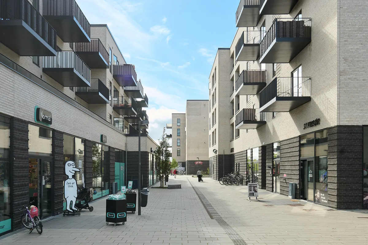Lejlighedsbygninger med balkoner og solpaneler og torve med butikker og stående cykler på Torvestrædet i Skovlunde. En person går med en babyvogn.