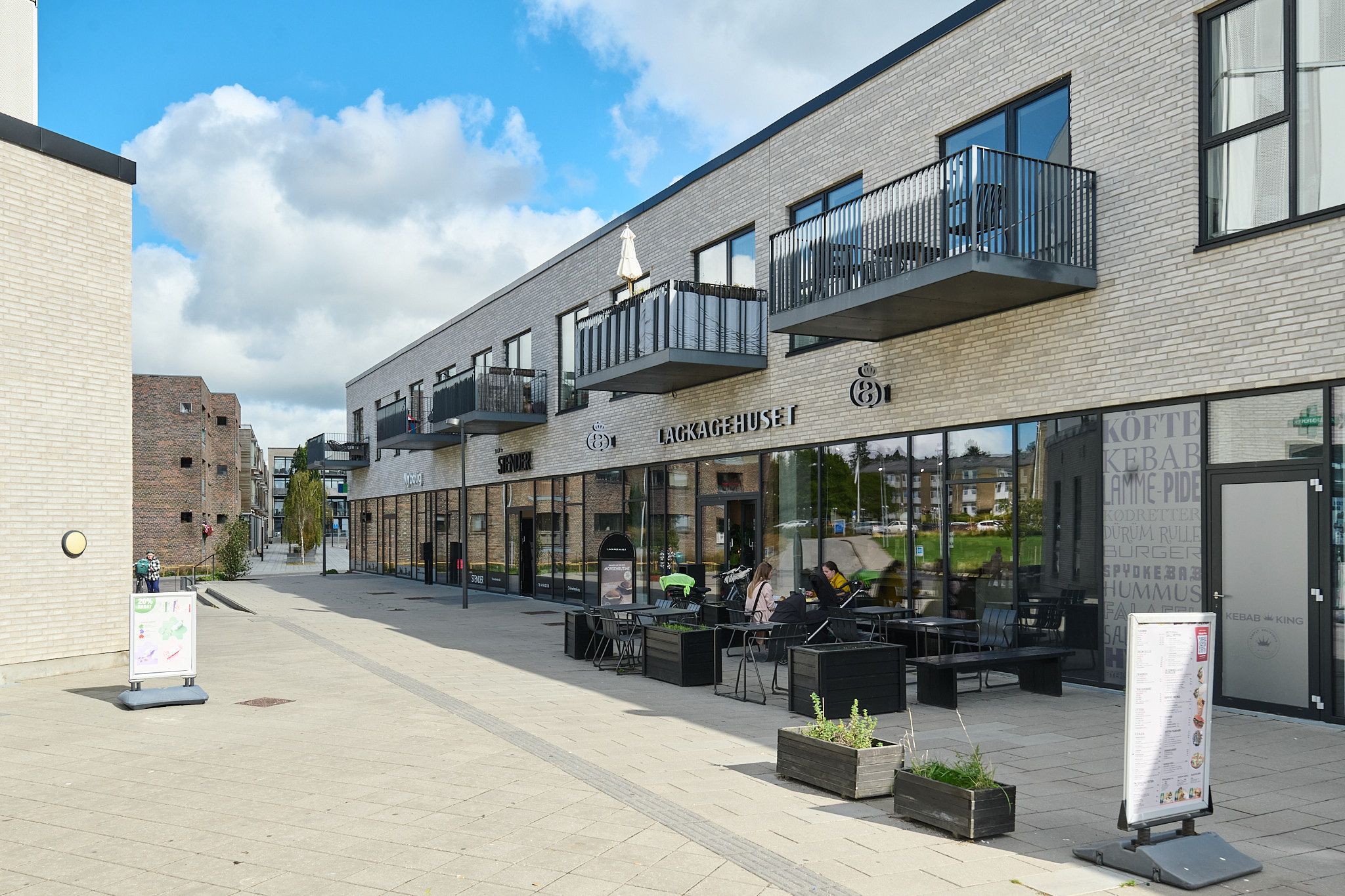 Moderne bygning med restaurant med terrasse, plantepot og menukort med tekst. En kvinde og en mand sidder og spiser.