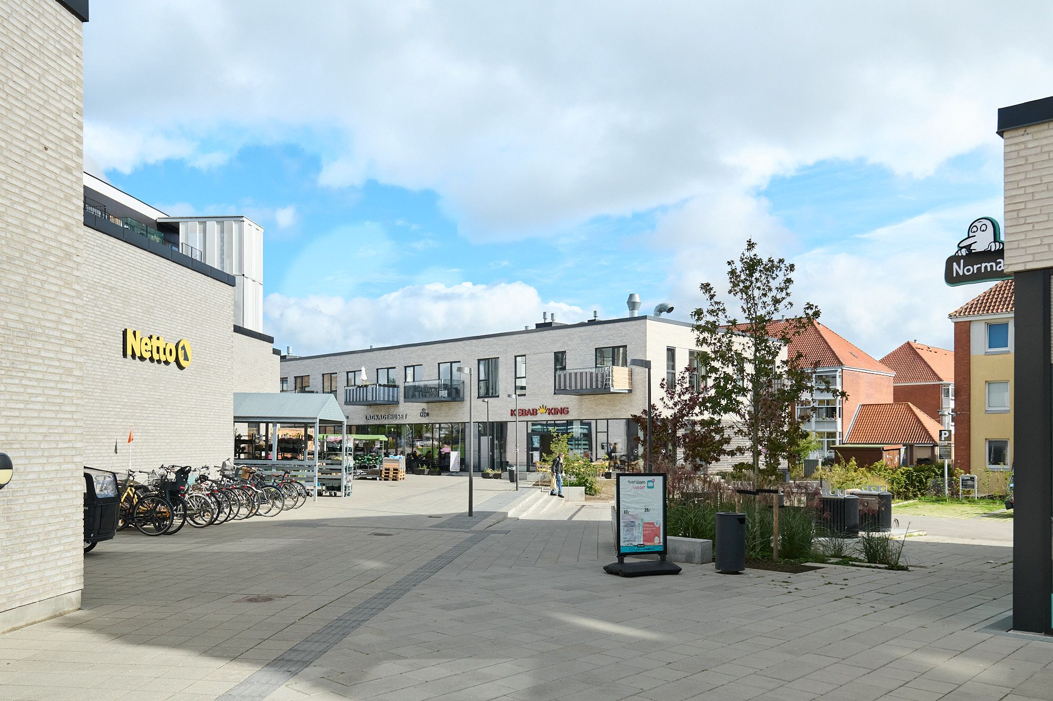 Lejemål med restaurant og takeaway muligheder. Bygning med Netto og Kebabs King. Parkerede cykler og vejsider. Sol og skyer på himlen.