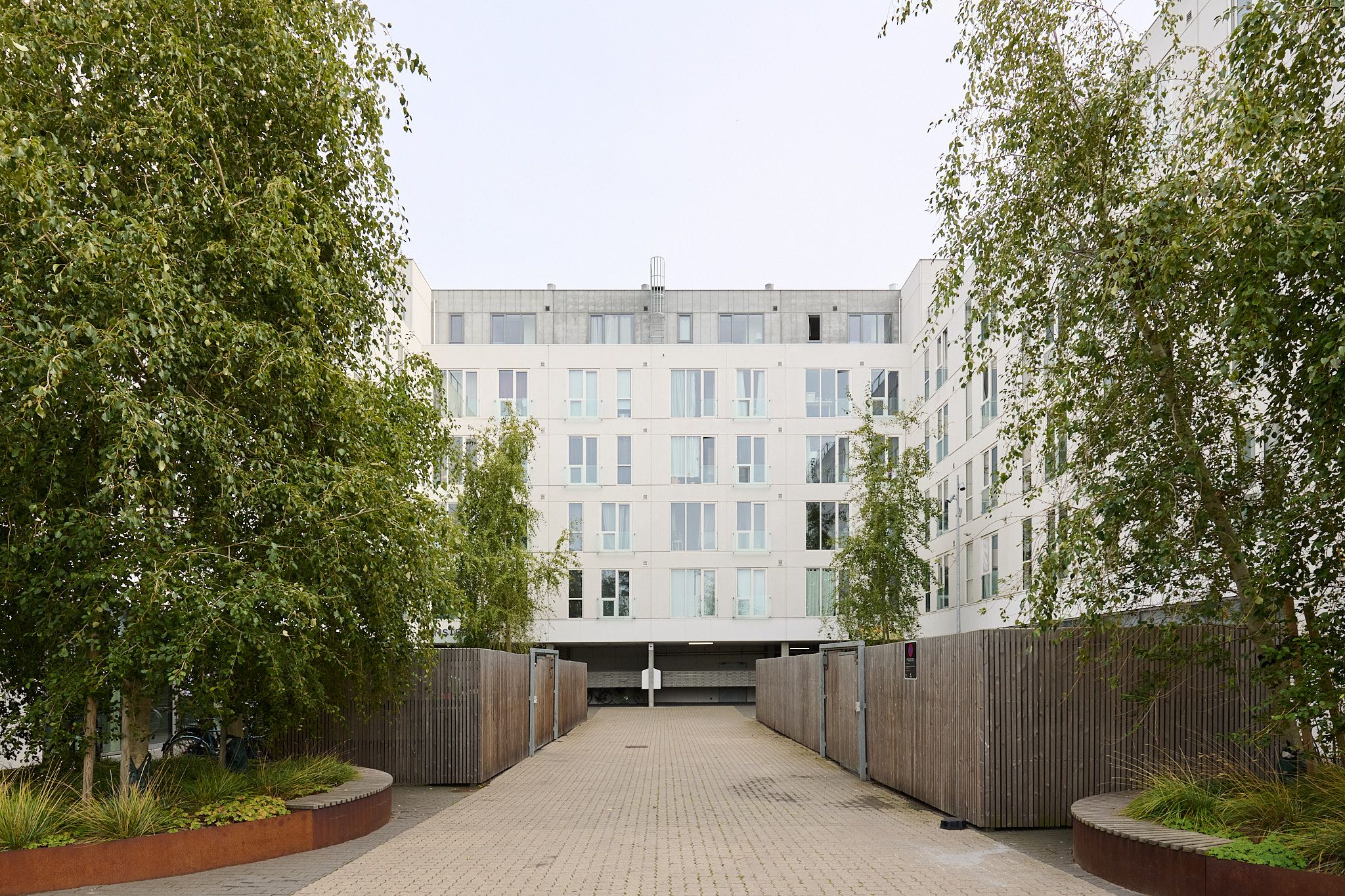 En moderne erhvervsbygning med glasvinduer og høje hvide vægge, omgivet af træhegn og træer. Stigende opgang med pavillon og skrånende vej mod en parkeringsplads