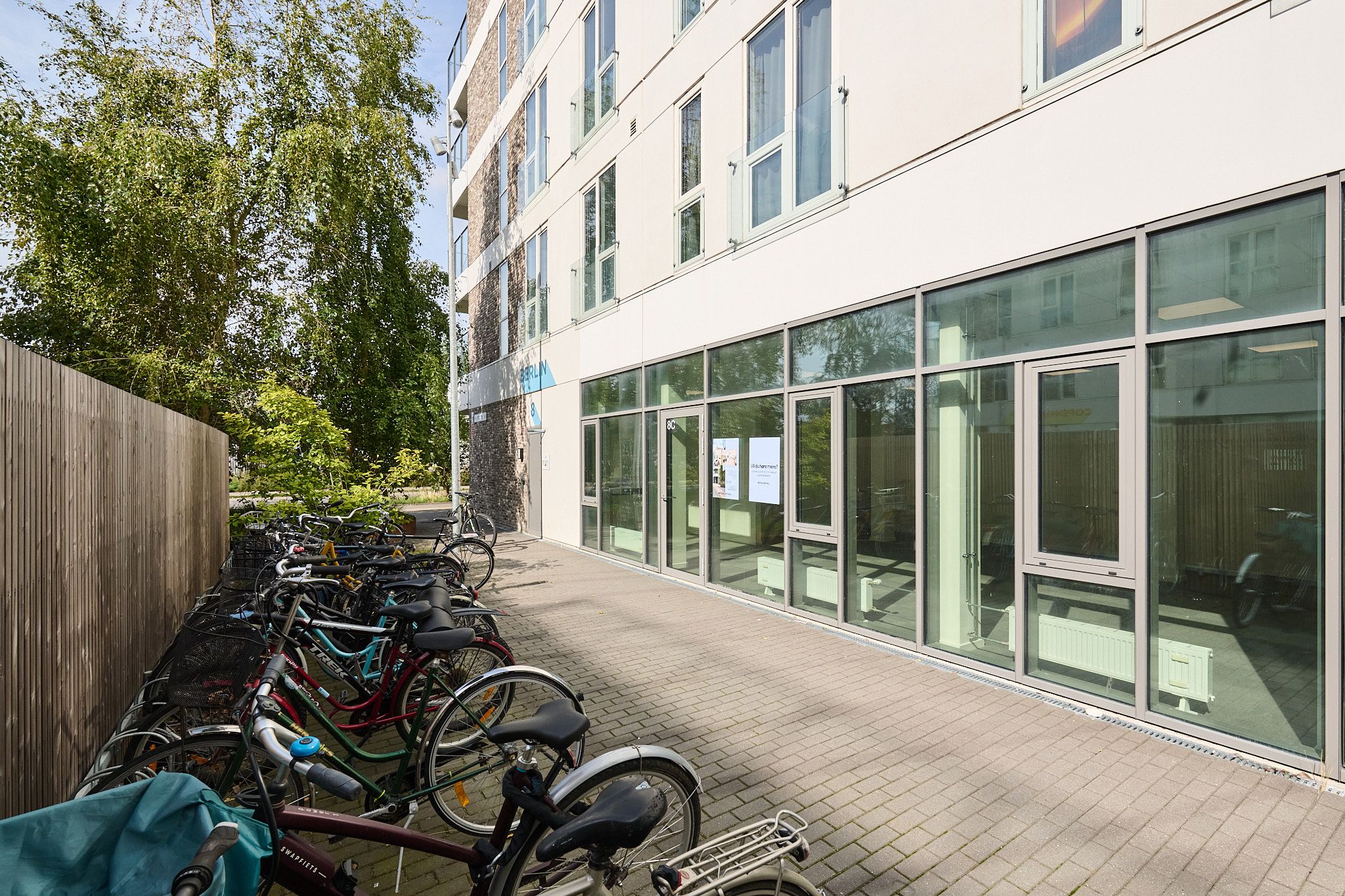 Flere cykler parkeret på siden af et hvidt bygningsfront med store ruder. Der er flere cykelparkeringspladser og et skur bagved.