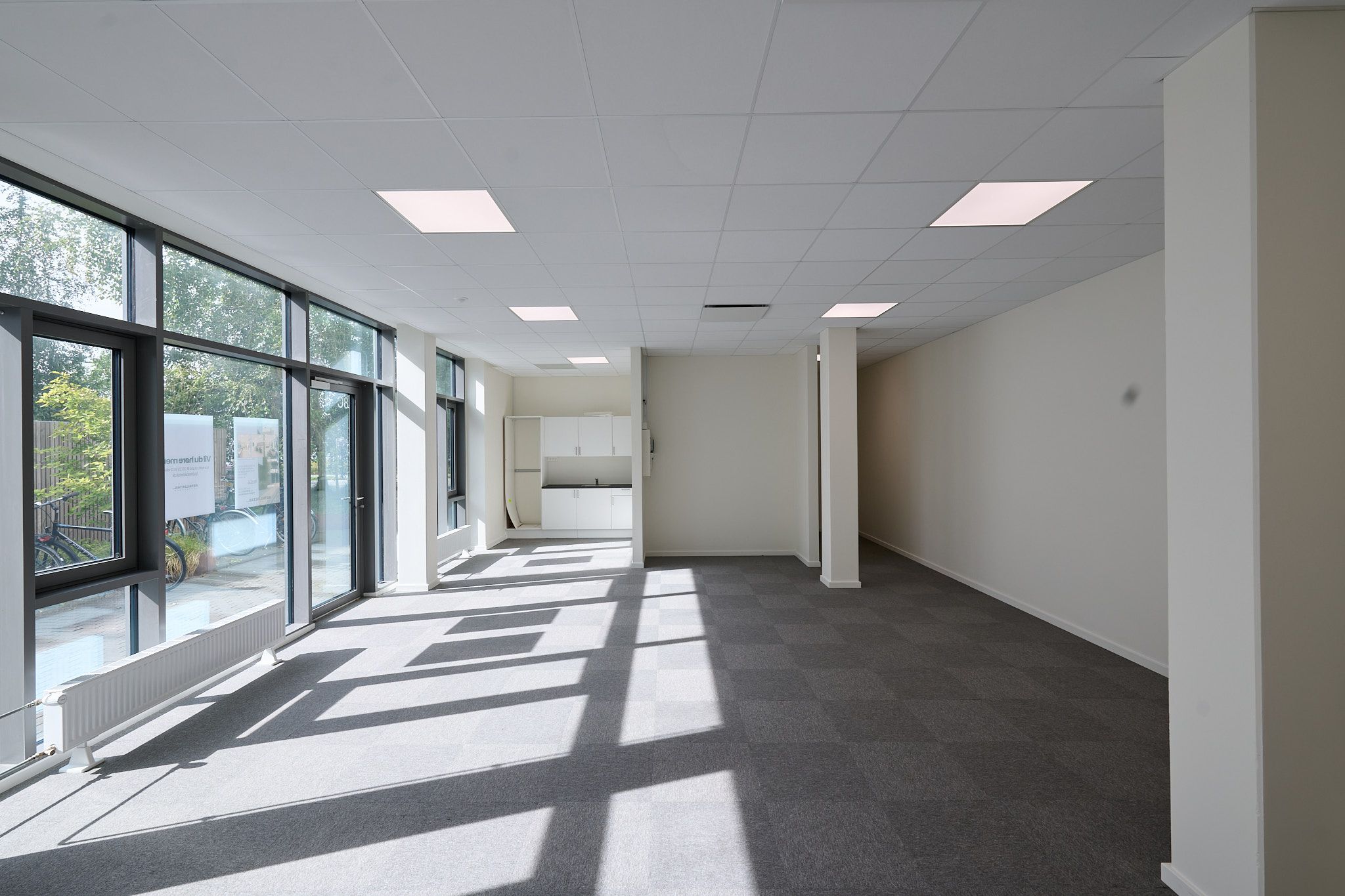 En tom lejlighed med lyse gulve, store ruder, hvide vægge og loft. Et køkken med hvide skabe og en hvid radiator. Skyggefulde mønstre på gulvet