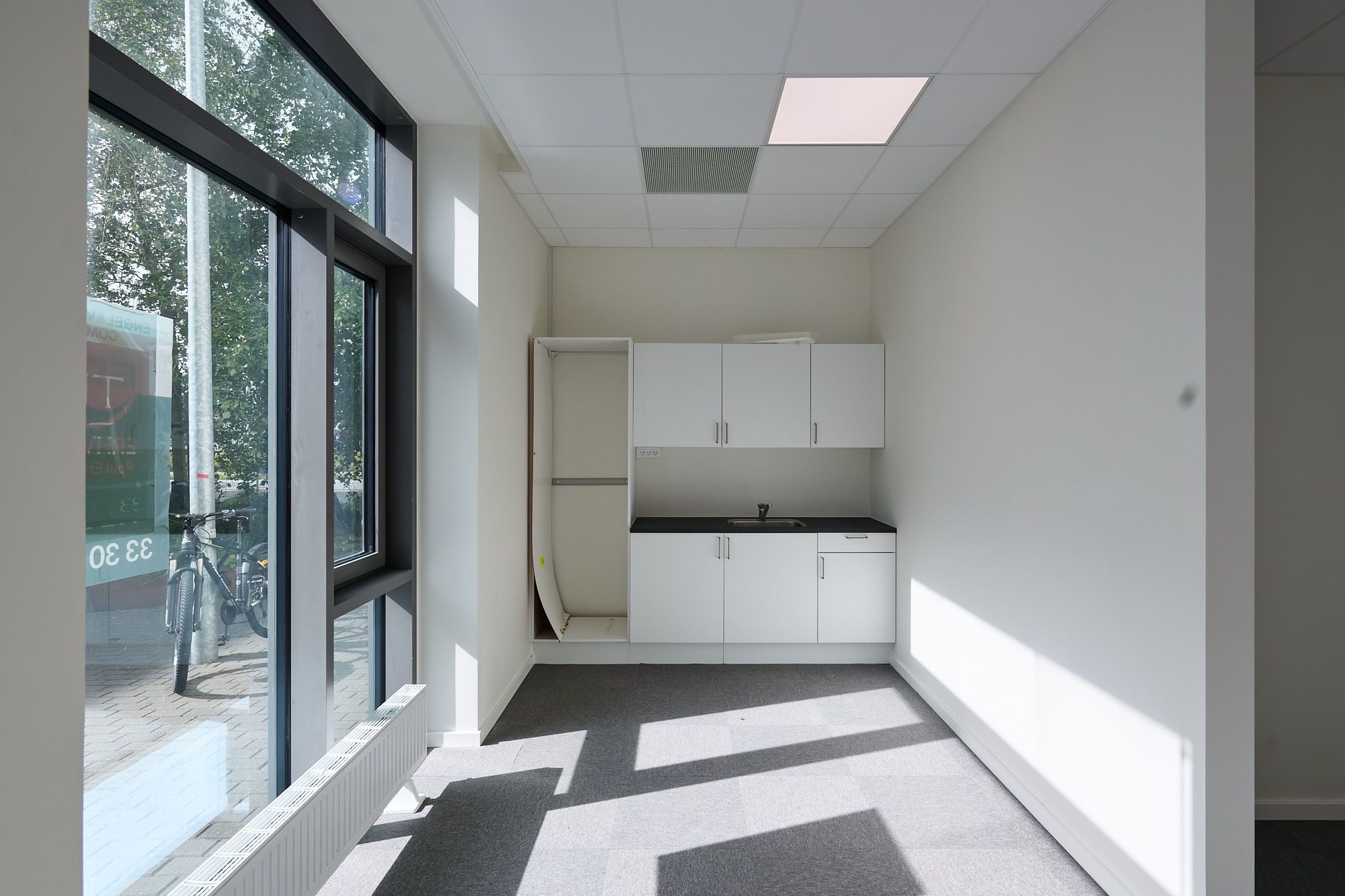 En lukket garderobe i hvidt, et køkken med hvide skabe og sort bordplade og en slank radiator i hvidt, placeret langs væggen i et kontorlokal