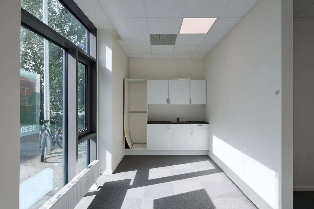 En lukket garderobe i hvidt, et køkken med hvide skabe og sort bordplade og en slank radiator i hvidt, placeret langs væggen i et kontorlokal