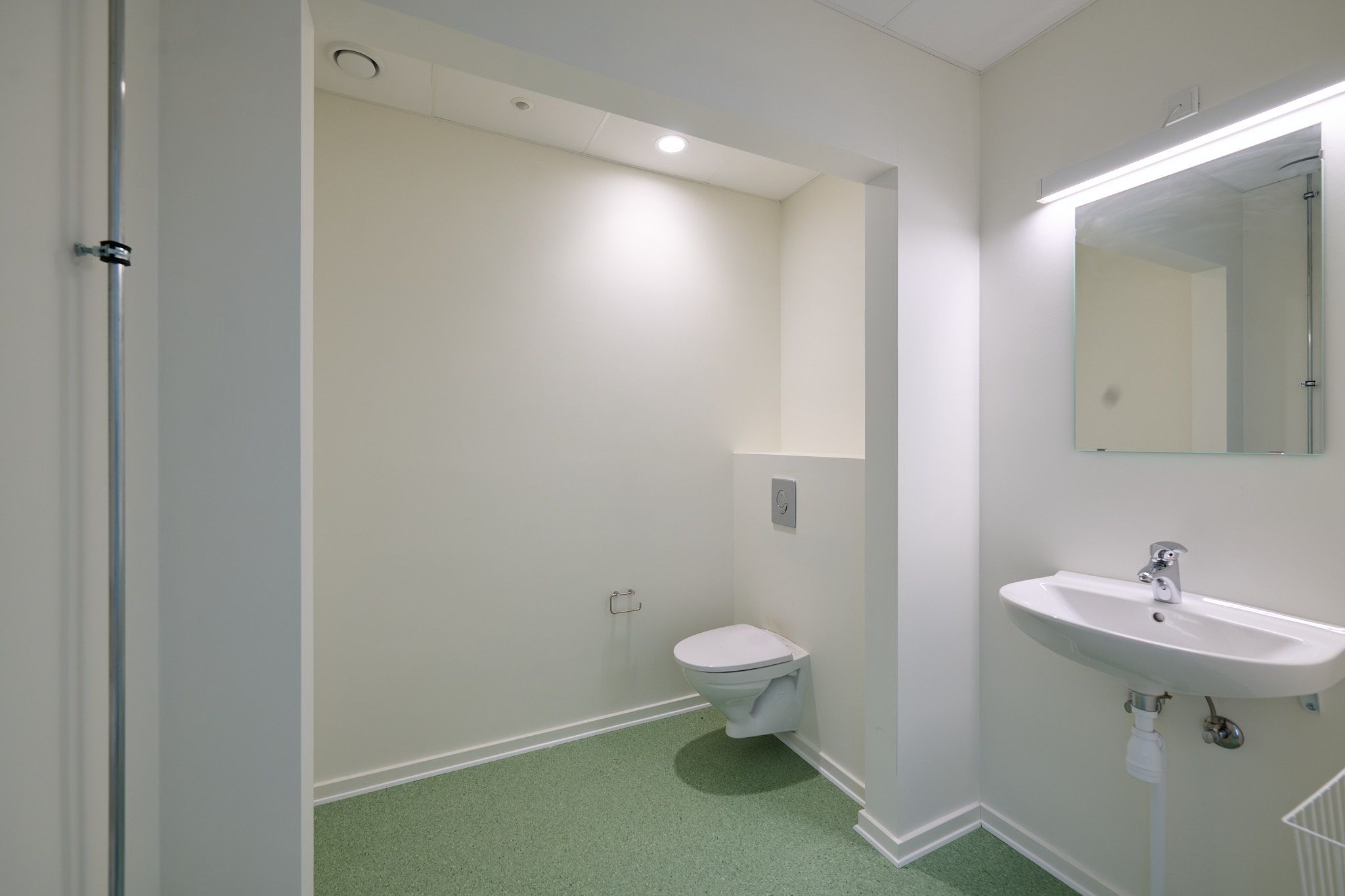 Et lille, hvidt badeværelse med en hvid toilet, hvid sink og en spejl. Der er et toiletpapirhylde ved siden af toilettet. Gulvet er grønt og væggene er hvide