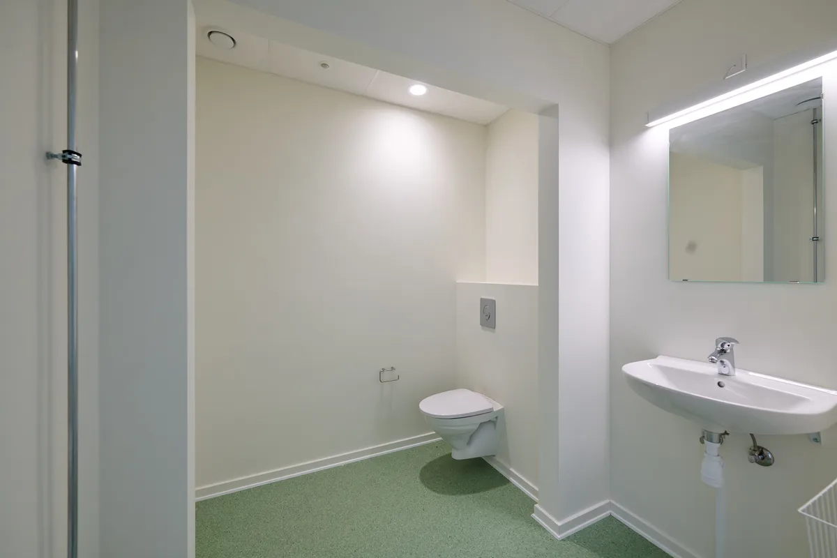 Et lille, hvidt badeværelse med en hvid toilet, hvid sink og en spejl. Der er et toiletpapirhylde ved siden af toilettet. Gulvet er grønt og væggene er hvide