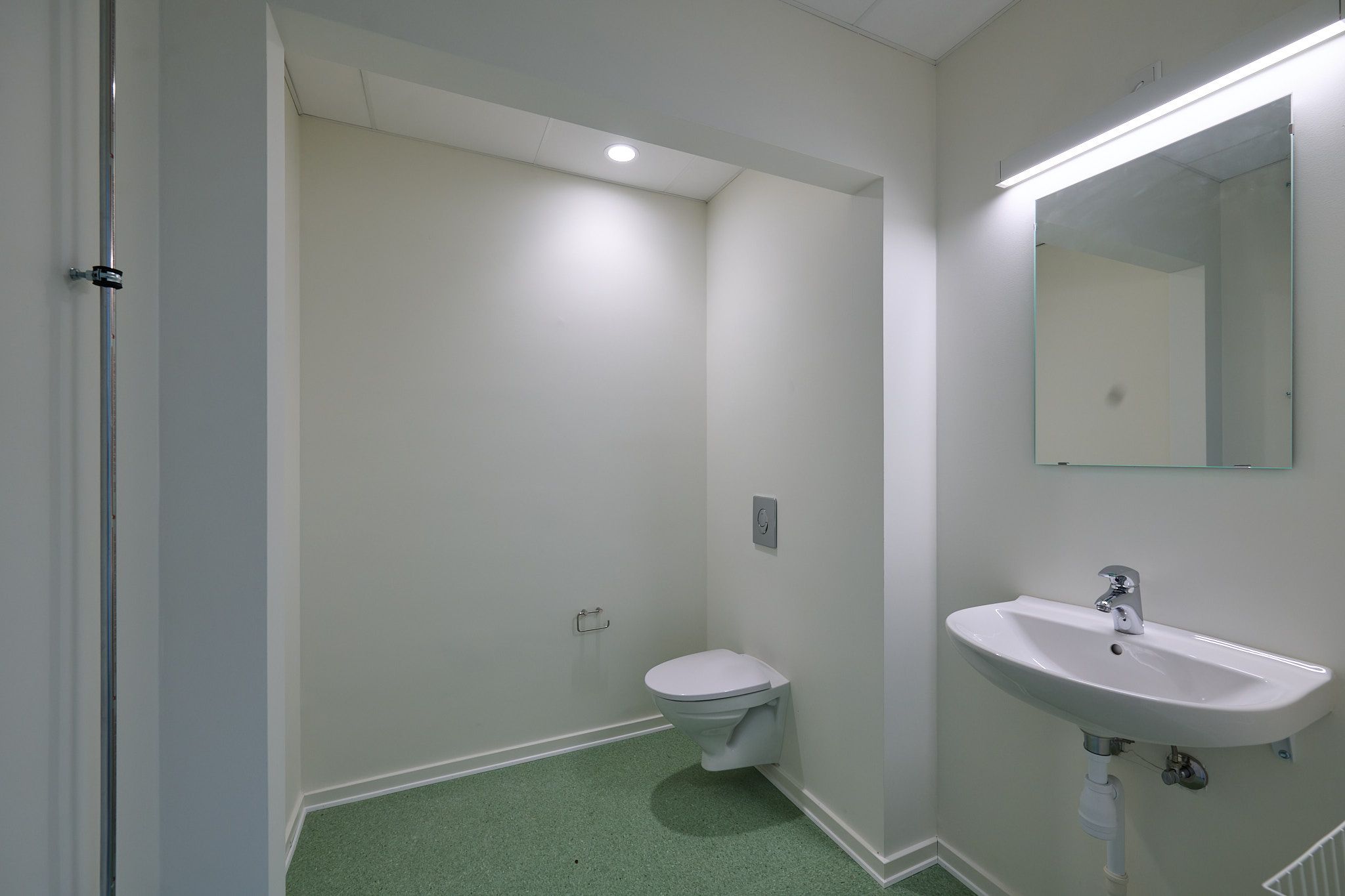 En lille, moderne toiletrum med hvidt toilet, hvidt vasketøj og grønt gulvtæppe samt en spejl med indbygget lys og et lys over toilettet