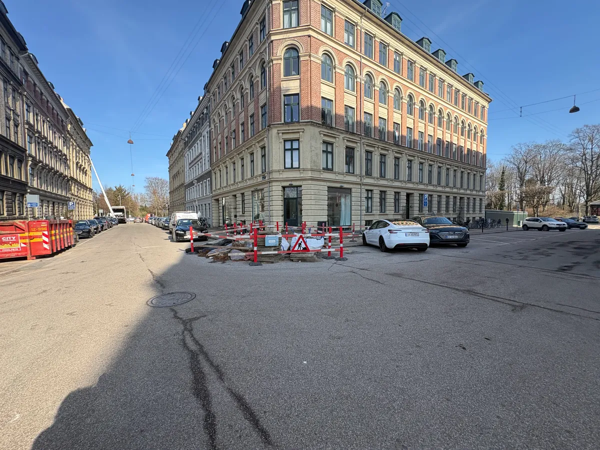 Store butikslokaler til leje på Olof Palmes Gade i København Ø. Bygning med flere etager og parkerede biler. Mange vinduer og højtrammer. Fortov og vej med røde bøsser og forhøjede fortove.