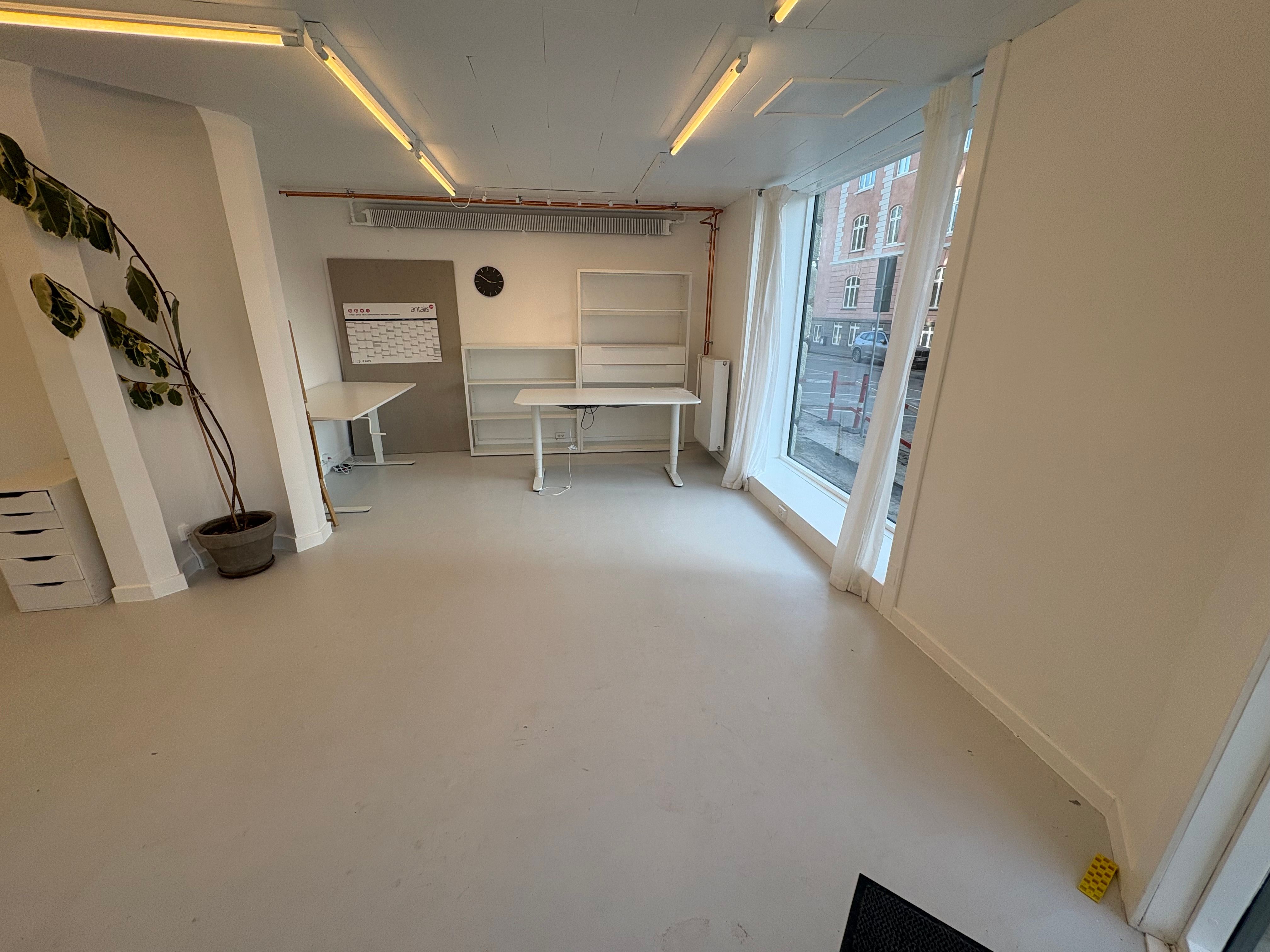 Foto af en tom og ren butik med hvidgulv og hvide vægge. Der er to hvide bord og højre er en højrehængs dør. Vindu med gardiner og skraldespand til højre.