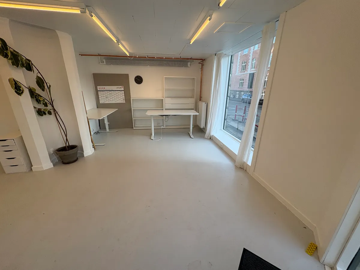 Foto af en tom og ren butik med hvidgulv og hvide vægge. Der er to hvide bord og højre er en højrehængs dør. Vindu med gardiner og skraldespand til højre.