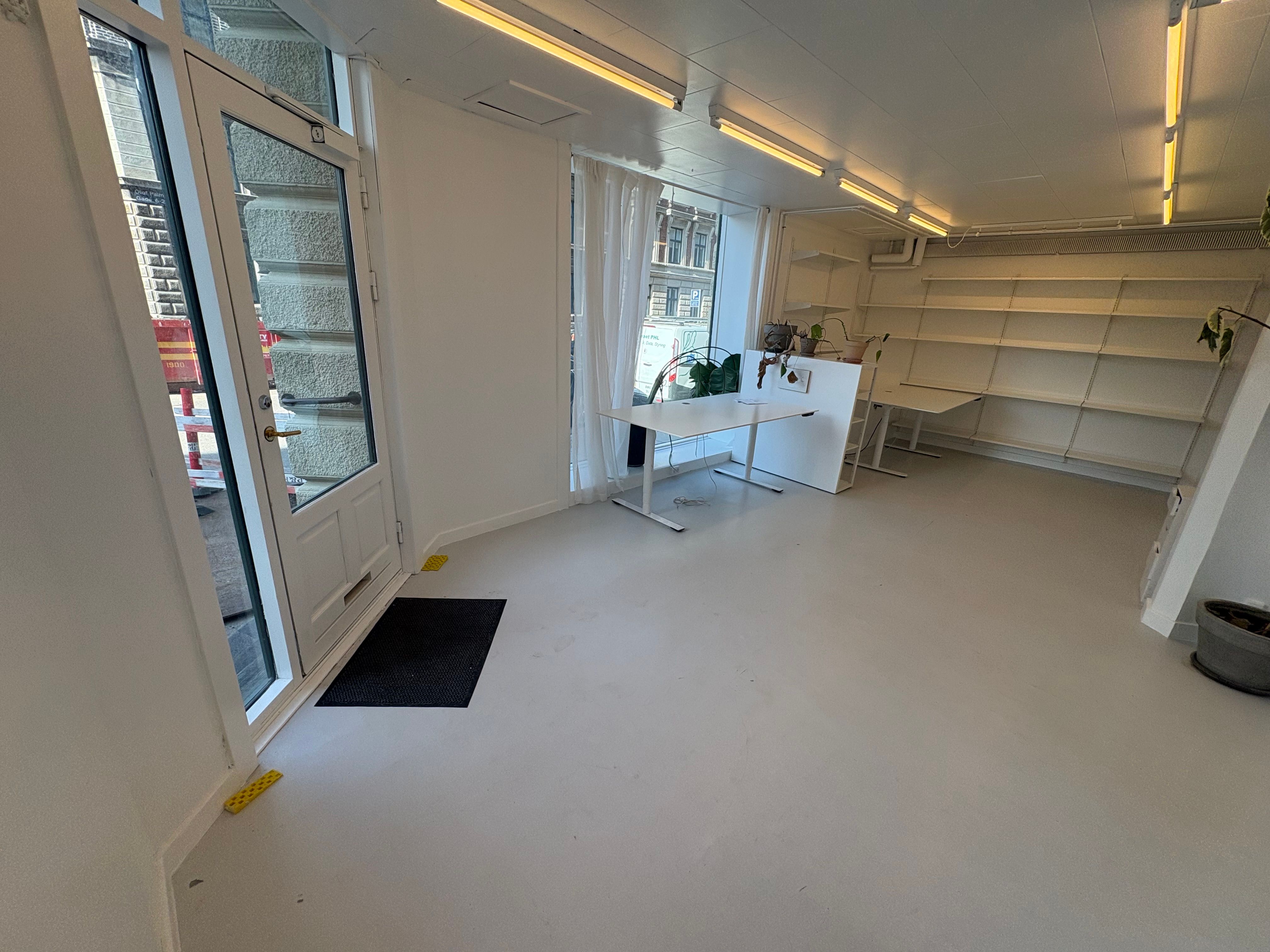 Et moderne kontor med hvide vægge og loft. To høje hvide skriveborde med højdejustering. Et lukket glasdør og en lukket gardin. To lyse, hvide hylder med læsestativ.