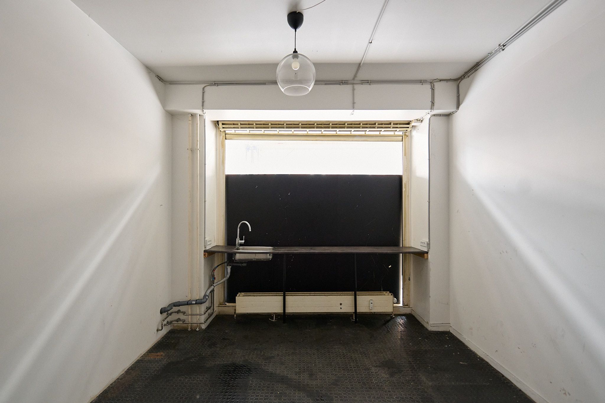 En tom kontorrum med lyse hvide vægge, sort gulv og en lysegrå loft. En enkelt skålvask er fastgjort til væggen og en enkelt lampe hænger fra loftet