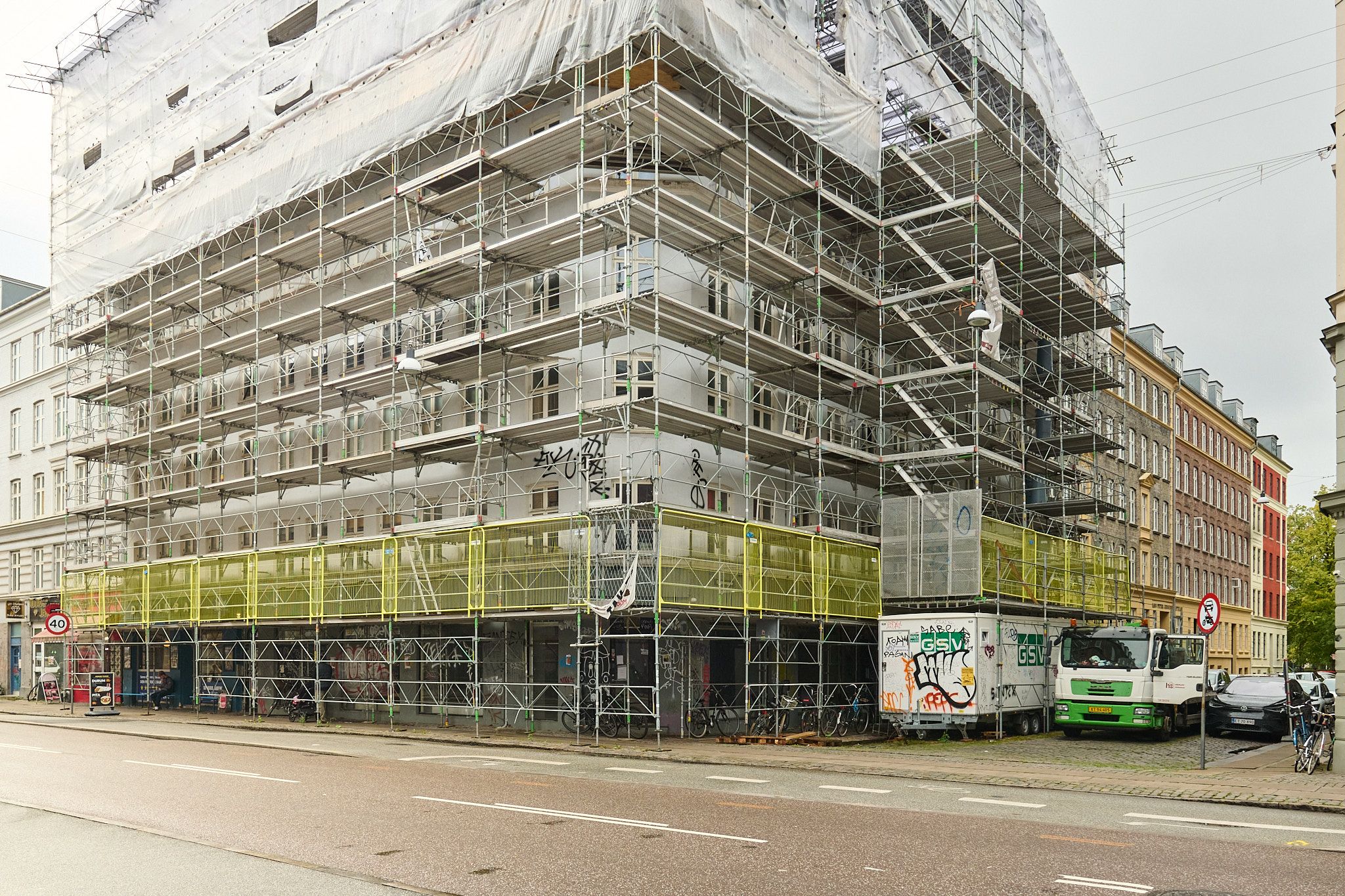 Byggested med stålskærme og stiger, røg fra en røgstige, to lastbiler parkeret på siden og to cykler på fortovet