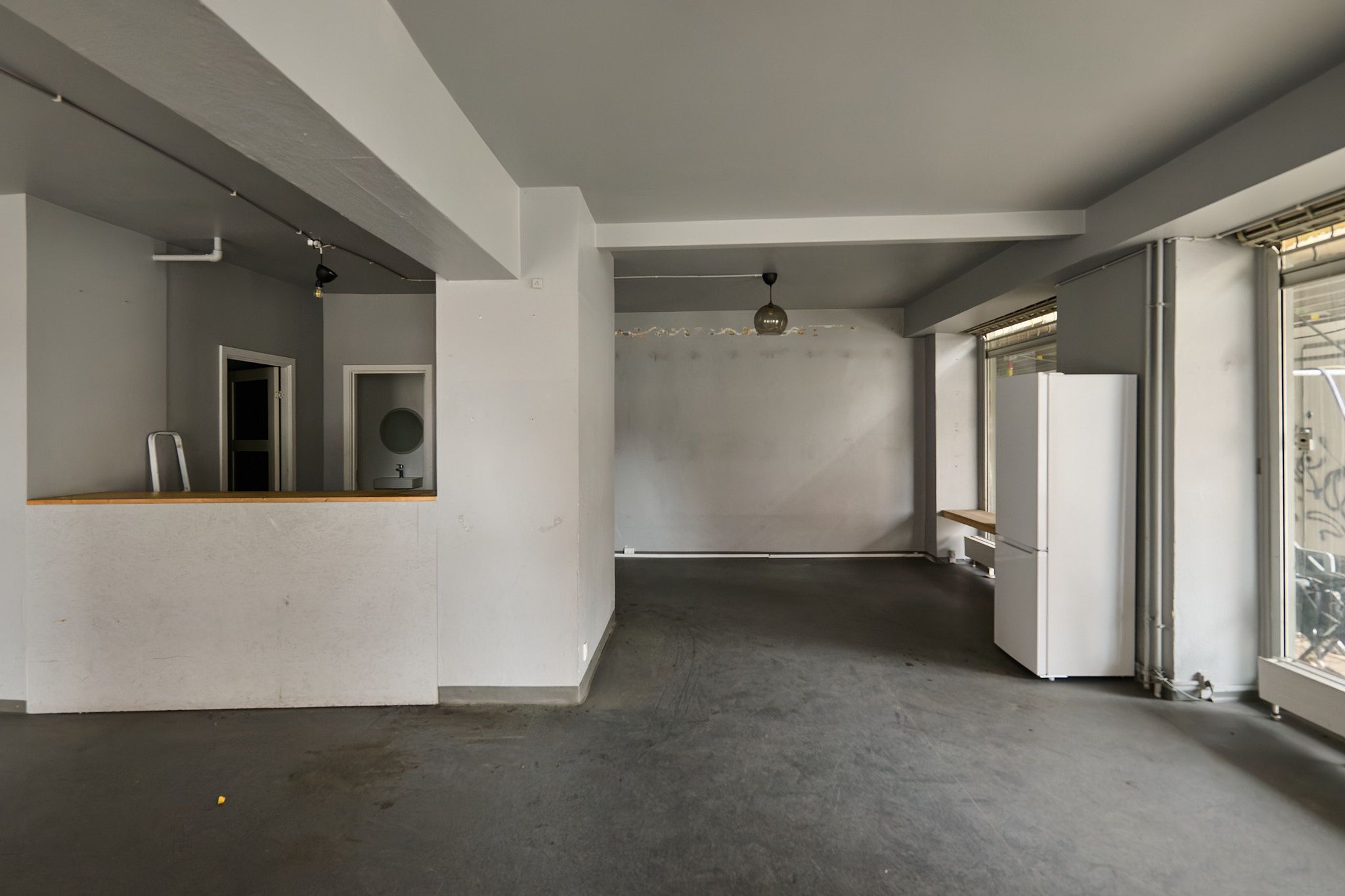 Rummeligt erhvervslejemål med lyse lofter og moderne interiør. Store vinduer og åben gulvplan. Et køleskab og en arbejdsbank er placeret. Lampehylde og loftslys giver god belysning.