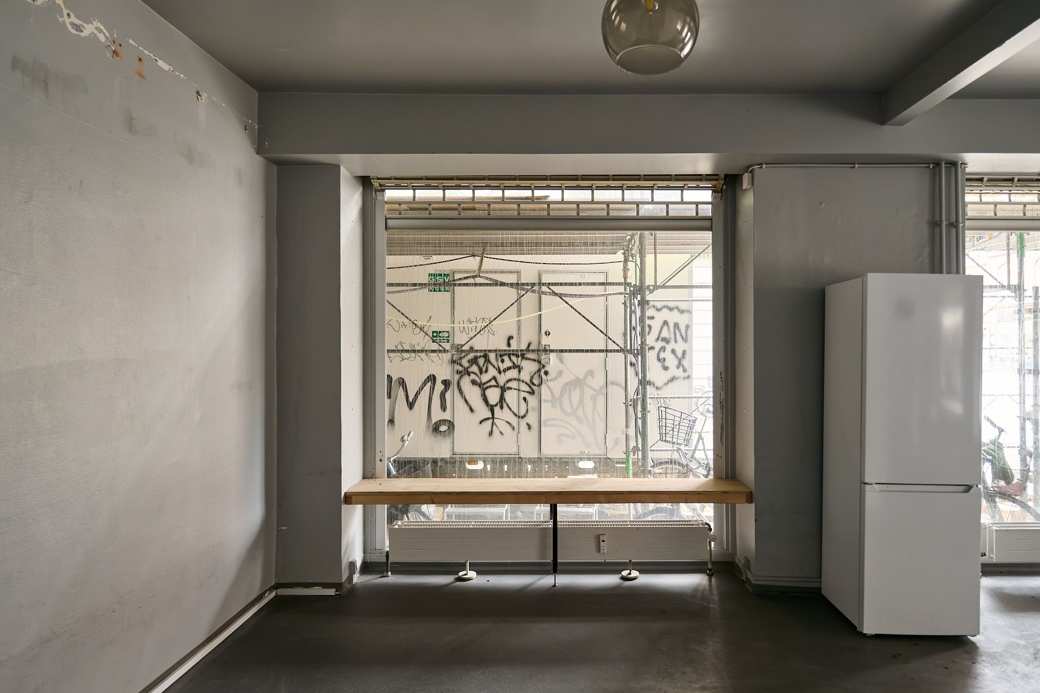 Moderne interiør med træbord og hvidt køleskab, lyse gulv og vægge samt store ruder med graffiti.