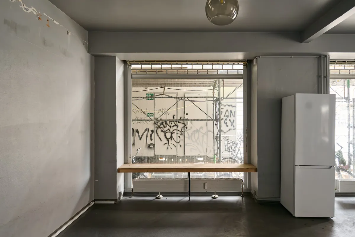 Moderne interiør med træbord og hvidt køleskab, lyse gulv og vægge samt store ruder med graffiti.