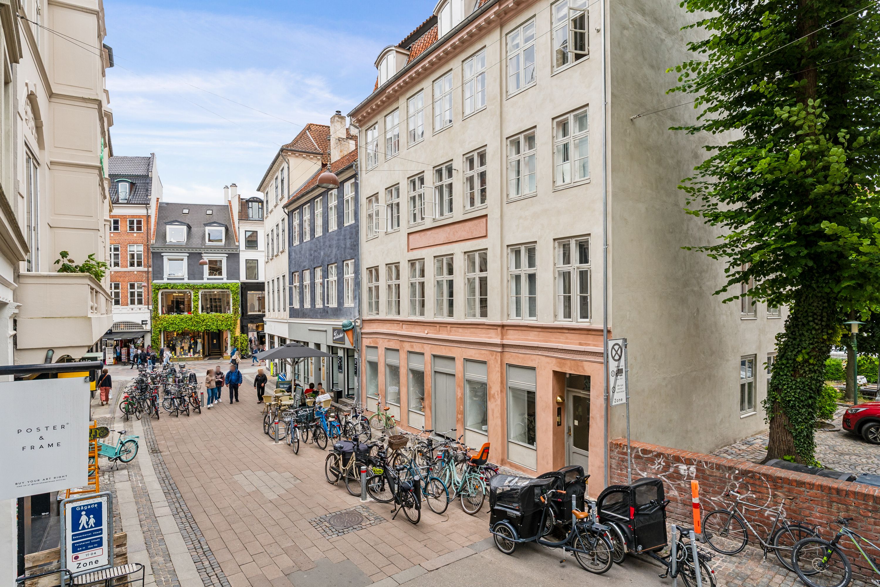 En cykelparkering med cykler og trehjulede vogne på Niels Hemmingsens Gade, mange bygninger og træer, samt gågade med mennesker og gadeudsmykning