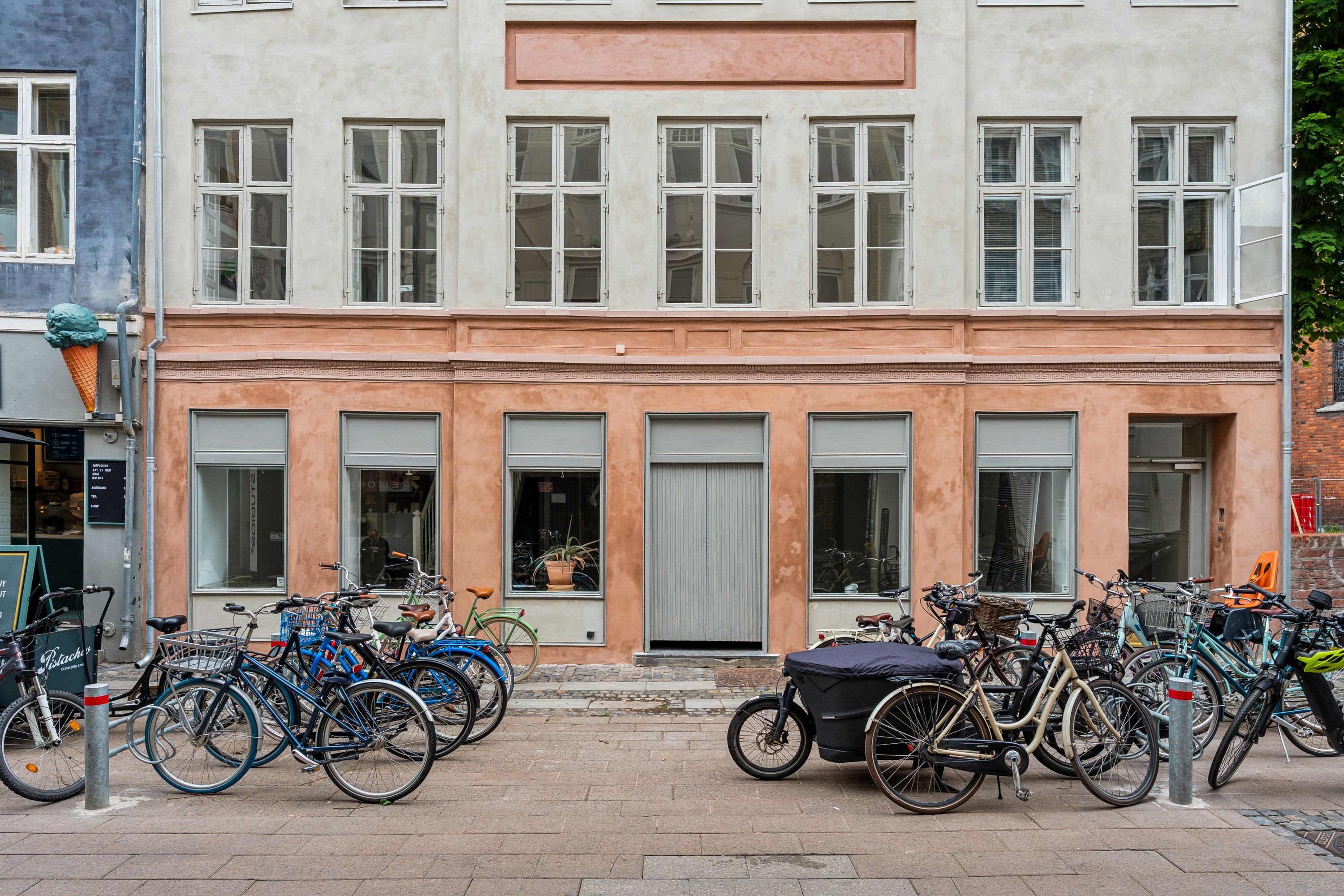Mange cykler er parkeret foran et forretningslokal med hvide ruder og en dekoration med en is, der er fastgjort til en rør. Lokalet er beliggende i et ældre bygningskompleks.