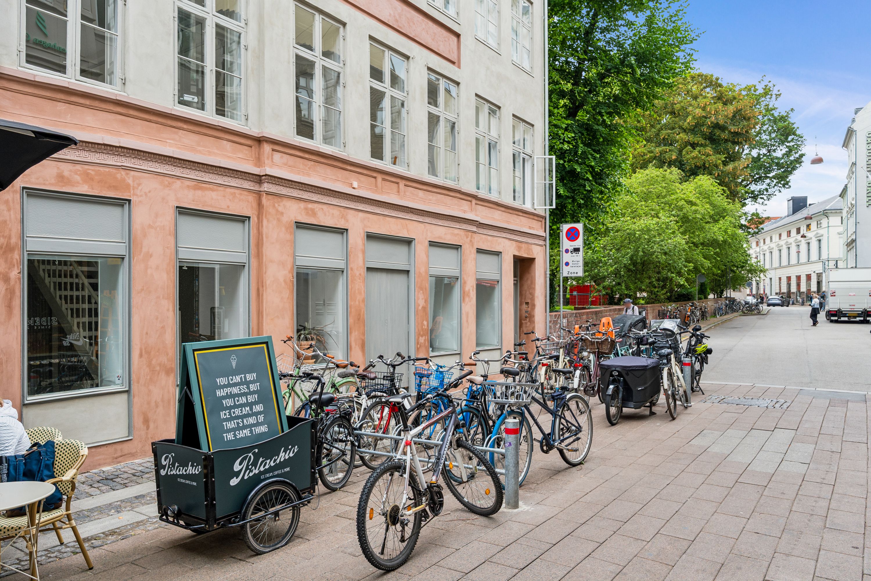 En række cykler står parkeret på en brolægget gade foran en bygning med en skilt med teksten Pistachio og en stol med en person.