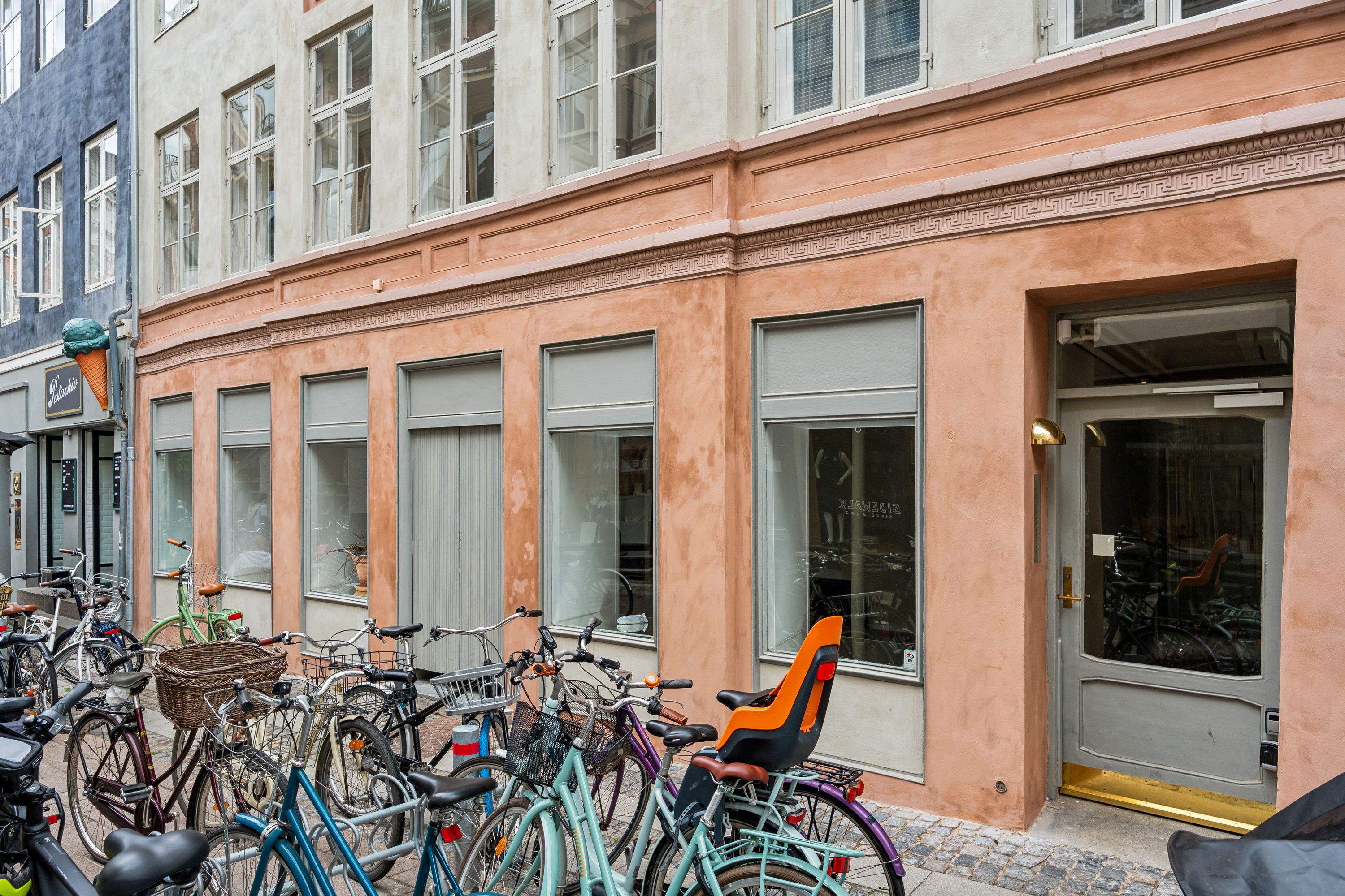 Butik i lysebrun bygning med møbelforretning, møbler og cykler parkeret udenfor og en isvogn i baggrunden
