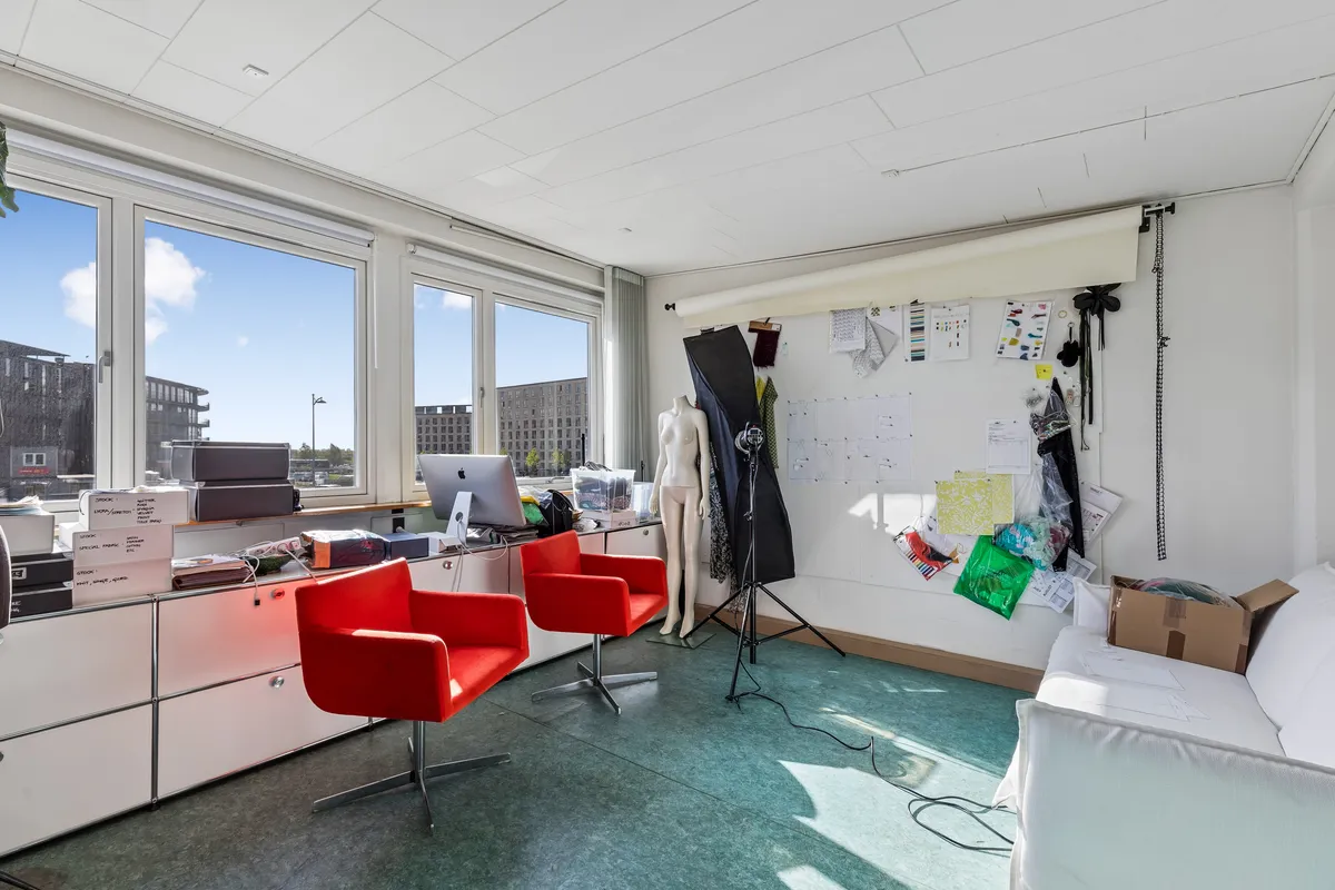 Kontor med skrivebord, computer, 2 røde stole, fotoudstyr og køkkensofa. Store vinduer og en udsigt til bygninger i baggrunden