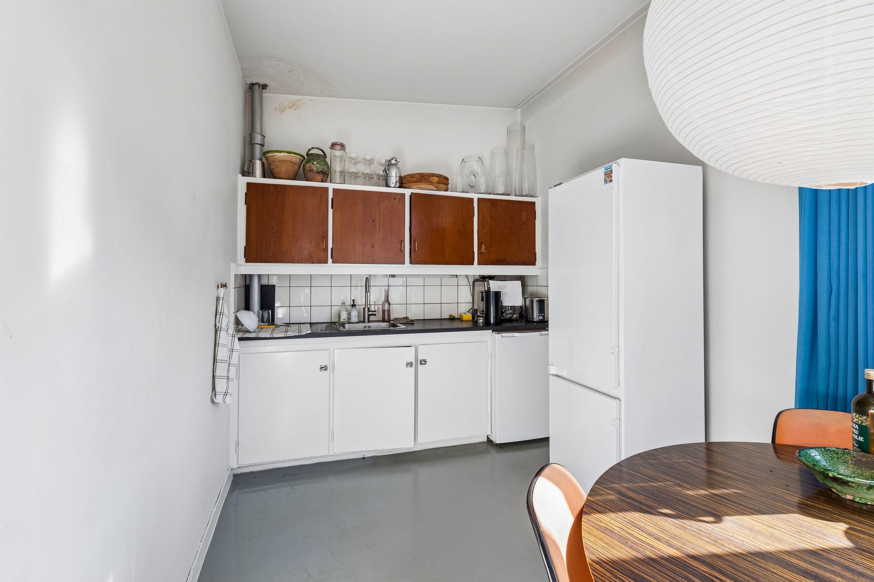En hvid køkken med lysegrå gulv, lysegrå loft, to hvide køleskabe og en rund hvid loftlampe med hvide lynlåg