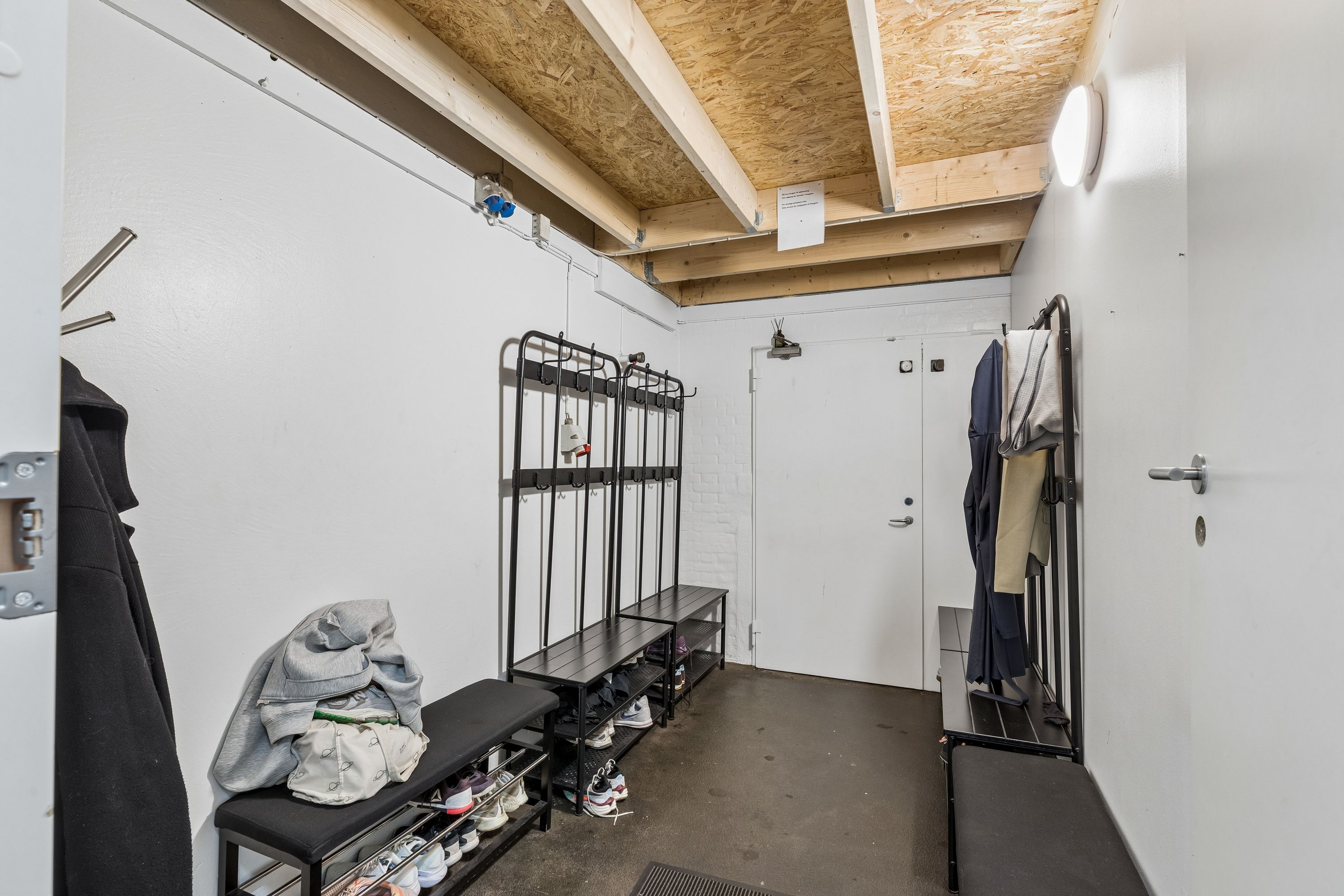 Indløbsområde med to tøjstativer og skohylderne. Hvide vægge med tøjhængere og to døre til højloftet lagerrum. Sorte sko, tøj og andre genstande liggende på hylderne