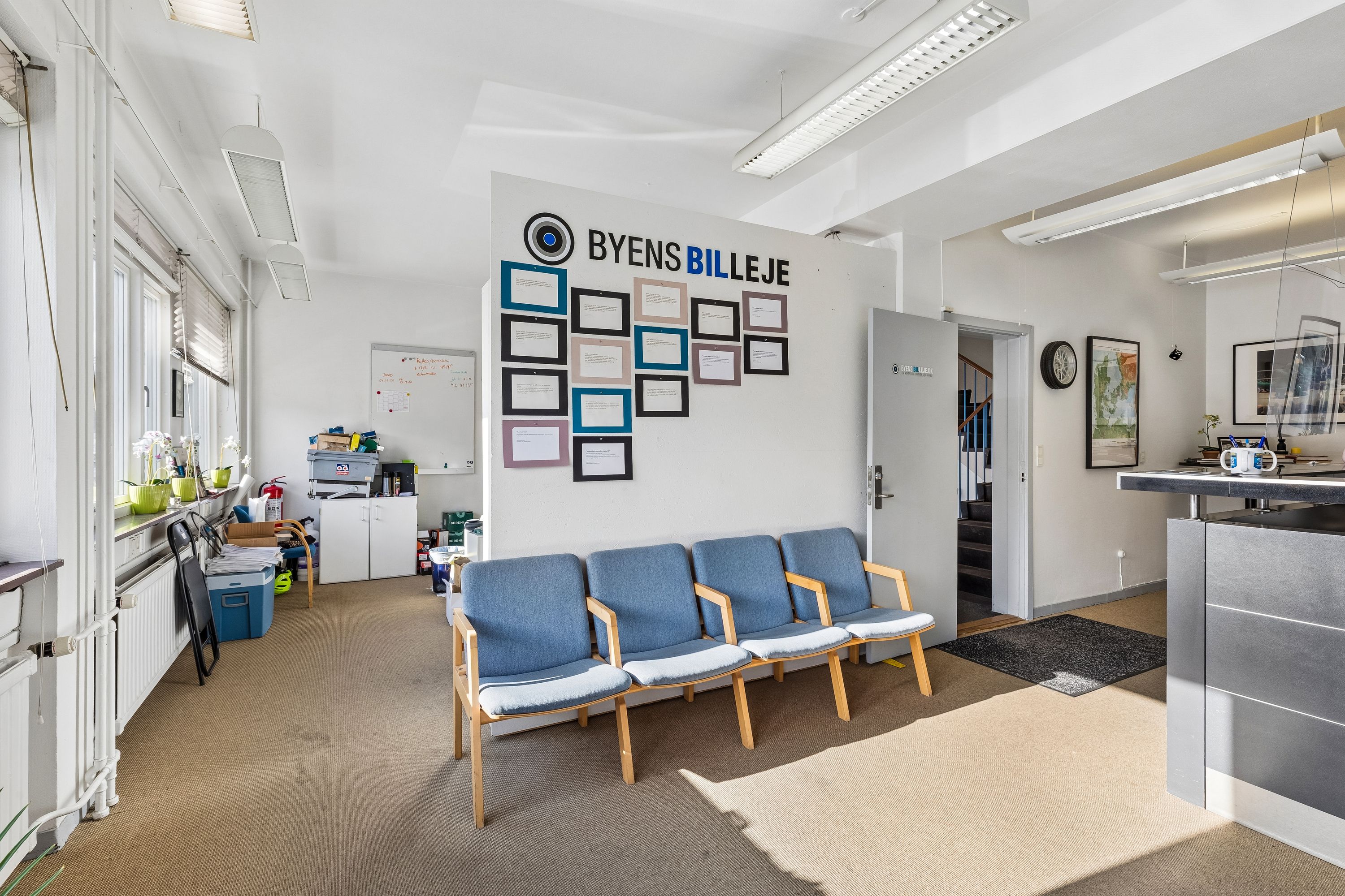 Kontorlokation med lyse loftsbelægninger og indvendige vægbilleder. Sofaer og høj skrivebord med flere personer i baggrunden. På væggen står "Byens Billeje" og en kalender.