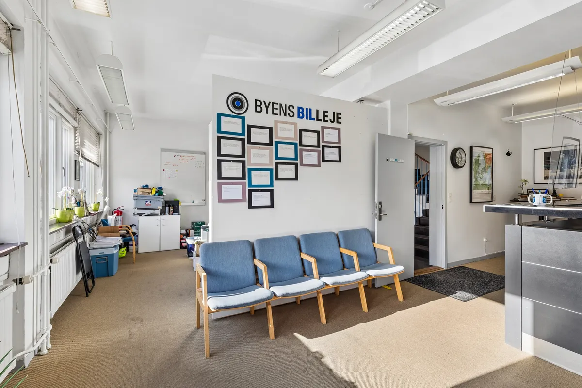 Kontorlokation med lyse loftsbelægninger og indvendige vægbilleder. Sofaer og høj skrivebord med flere personer i baggrunden. På væggen står "Byens Billeje" og en kalender.