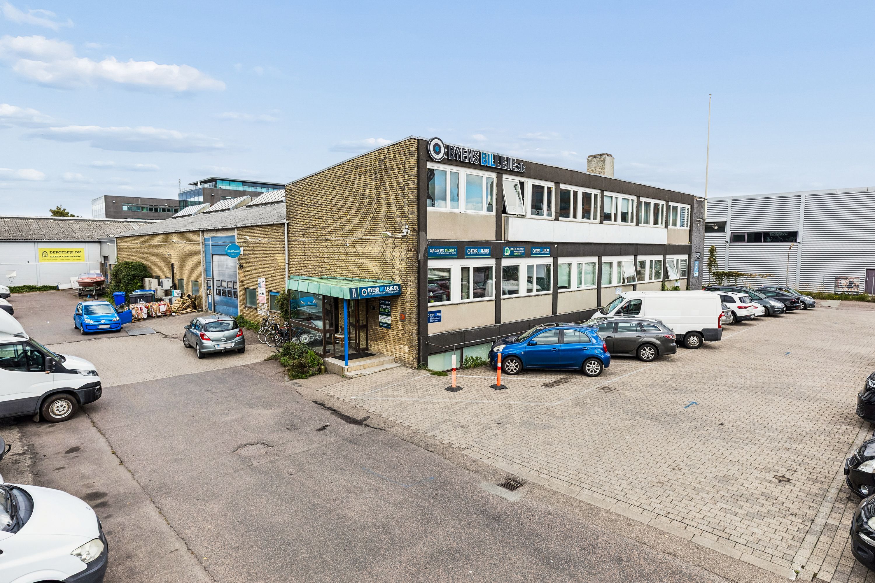 Flere biler parkeret på Bådehavnsgade i København. Kontorbyggeri med skrift Byens Billeje.dk. Solrig himmel med skyer. Stiger med højt fremspring.
